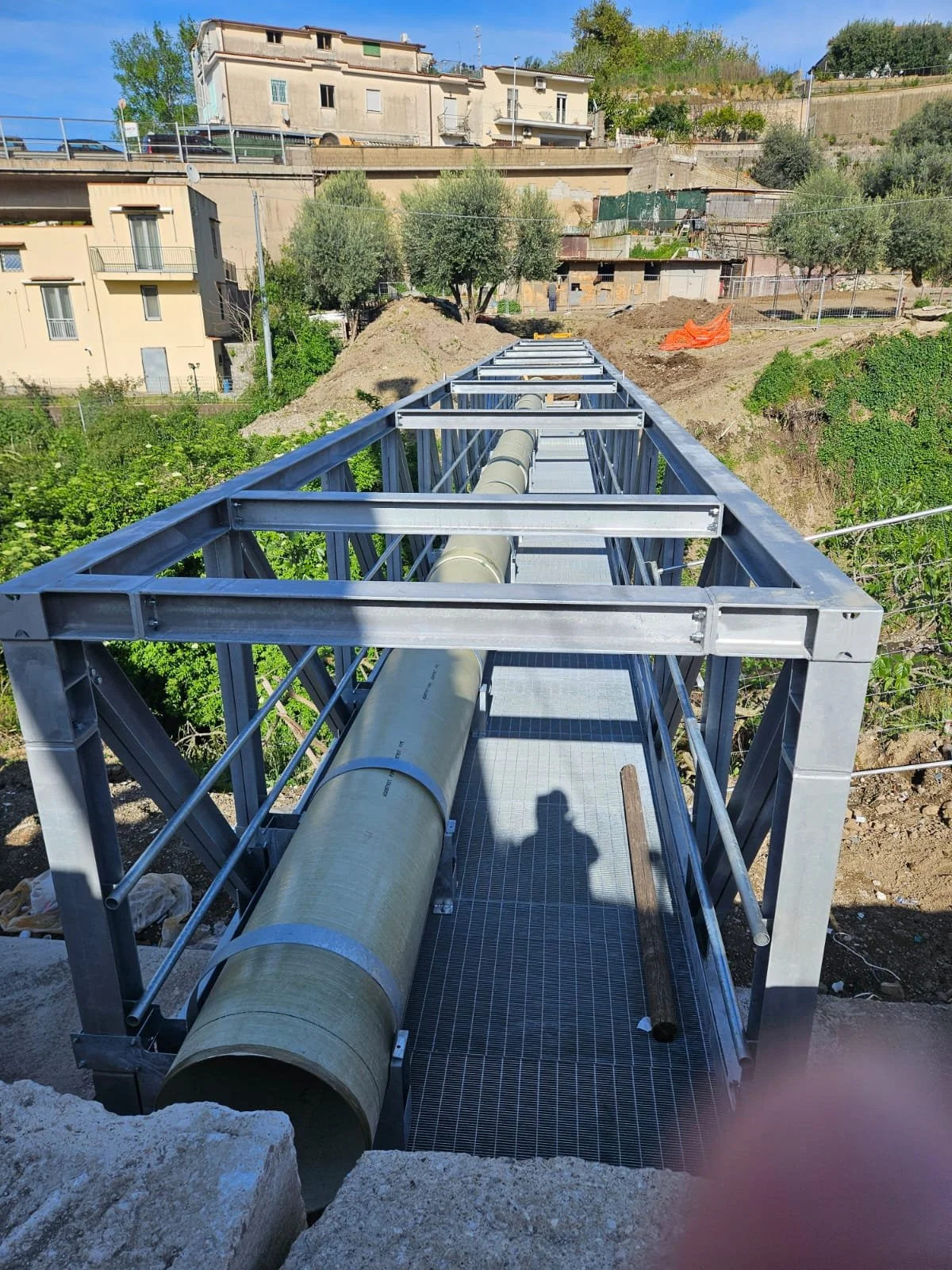 Struttura metallica in fase di installazione con un grande tubo di cemento disposto lungo la passerella, sullo sfondo case e vegetazione in un'area in salita.