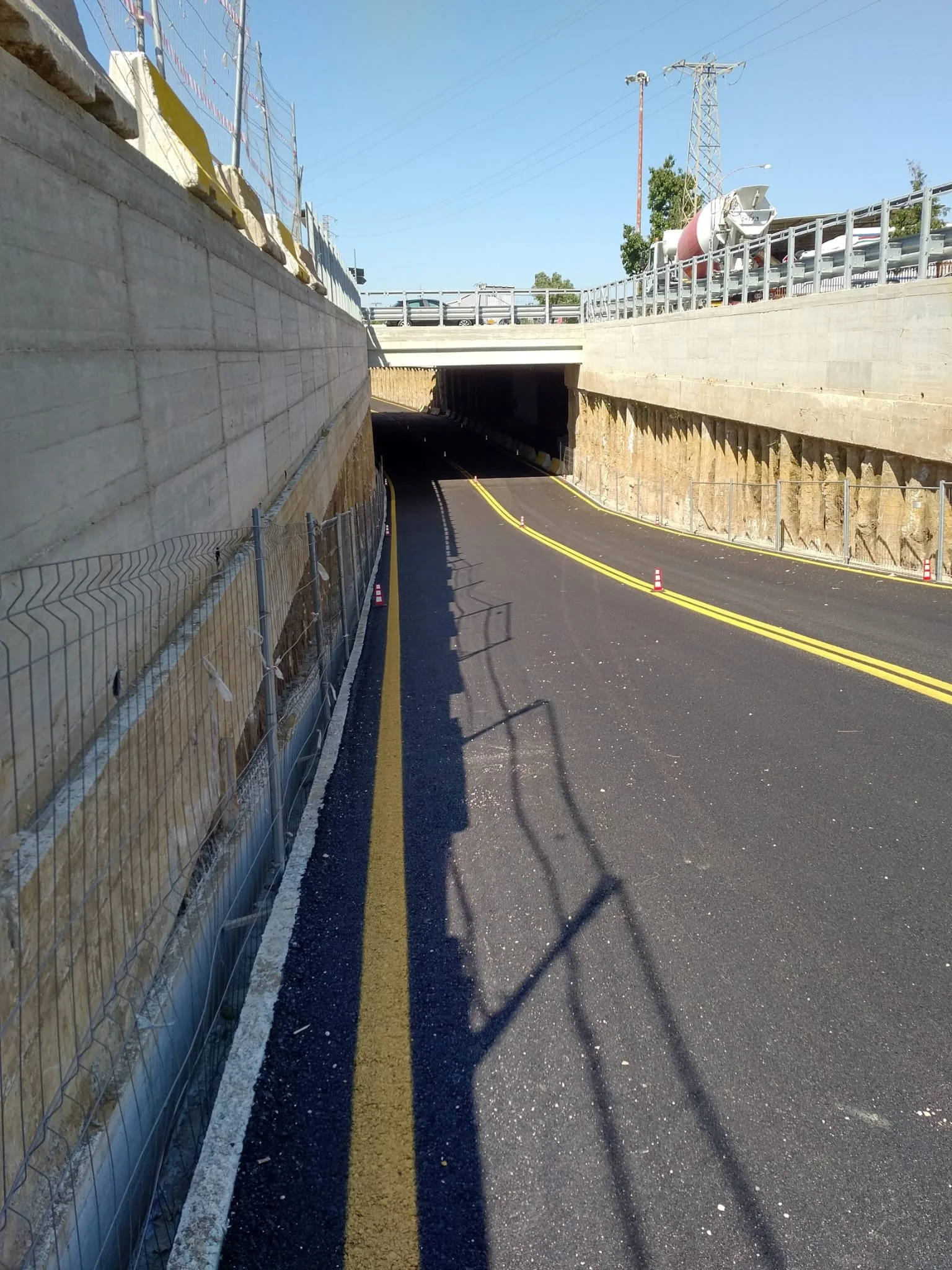 Strada di montagna con galleria, tagliata nella roccia, con linee di separazione gialle e ostacoli di traffico, sotto un ponte, con infrastrutture e cavi sopra.