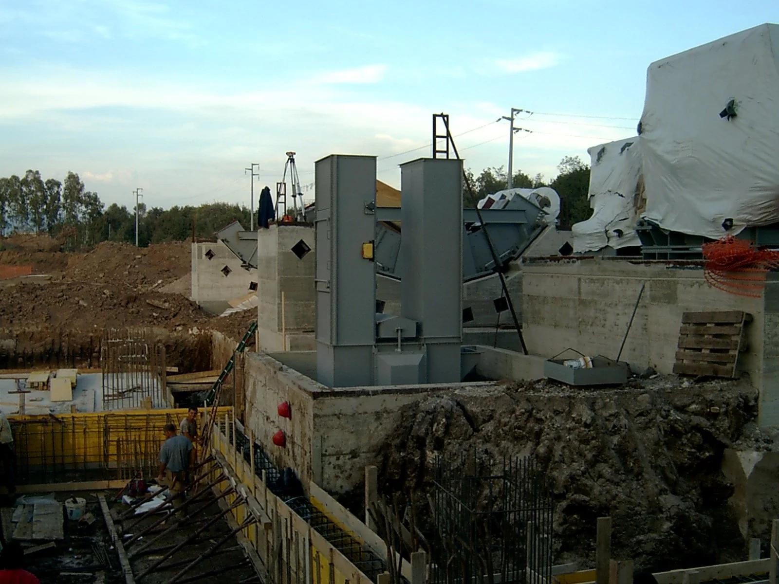 Cantiere edile con lavori in corso, ponteggi e strutture temporanee, terreno scavato, pochi operai al lavoro.