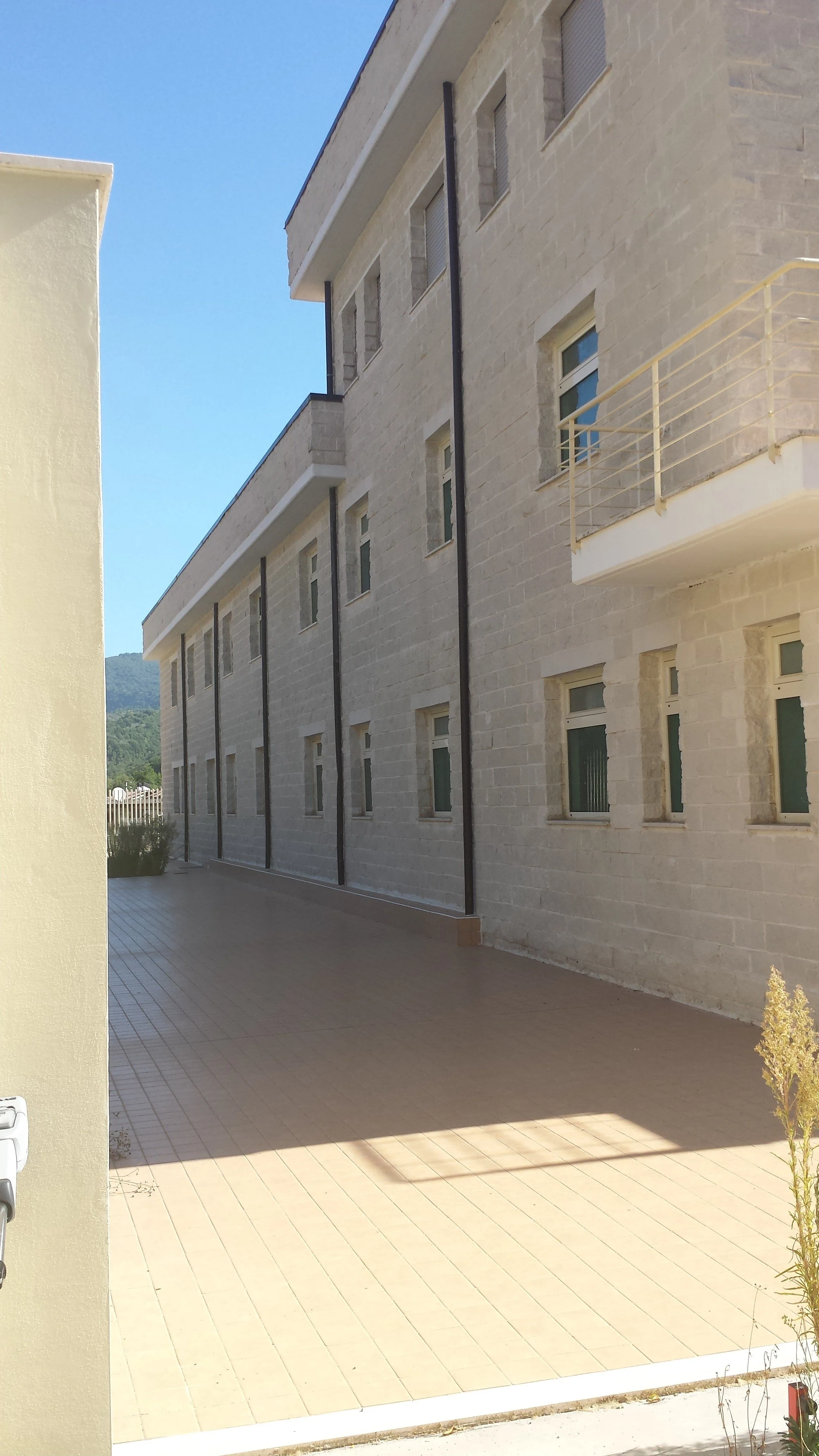 Fotografia di un edificio con pareti in pietra e più finestre, vista da un cortile aperto con piastrelle beige e un'ombra. Sullo sfondo si vede una collina verde.