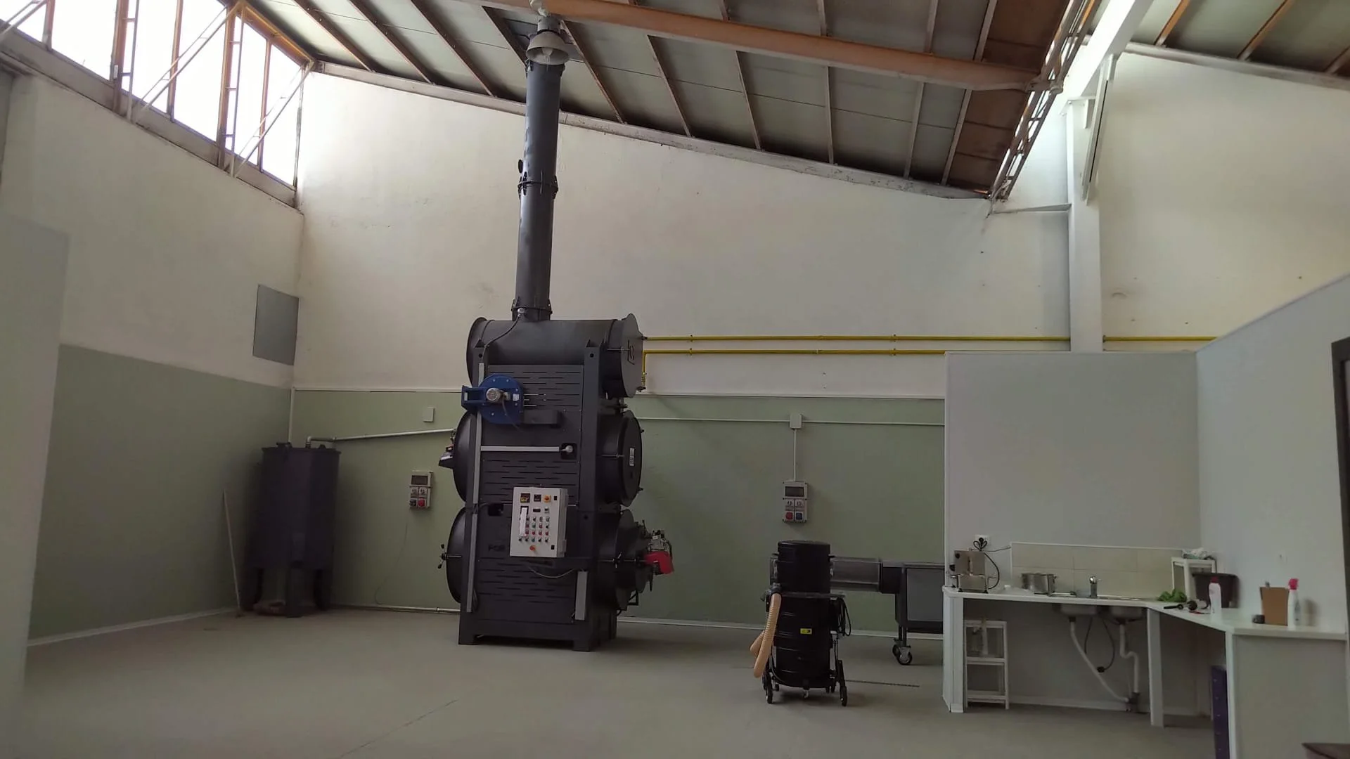 Immagine di una camera di ricerca o laboratorio con una grande apparecchiatura cilindrica nera, probabilmente un'unità di combustione o di riscaldamento, con un tubo di scarico che sale verso il soffitto alto. Sul lato sinistro ci sono due grandi ser