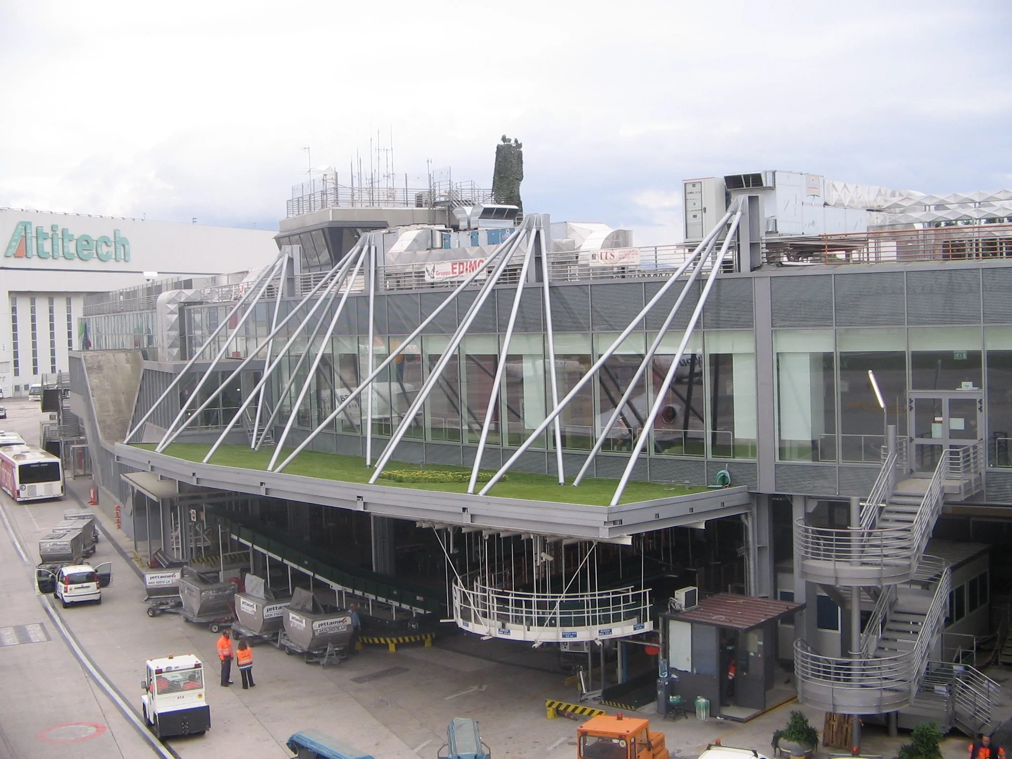 Immagine di un terminal aeroportuale moderno con strutture di vetro e metallo, alcune persone e veicoli vicino all'edificio.