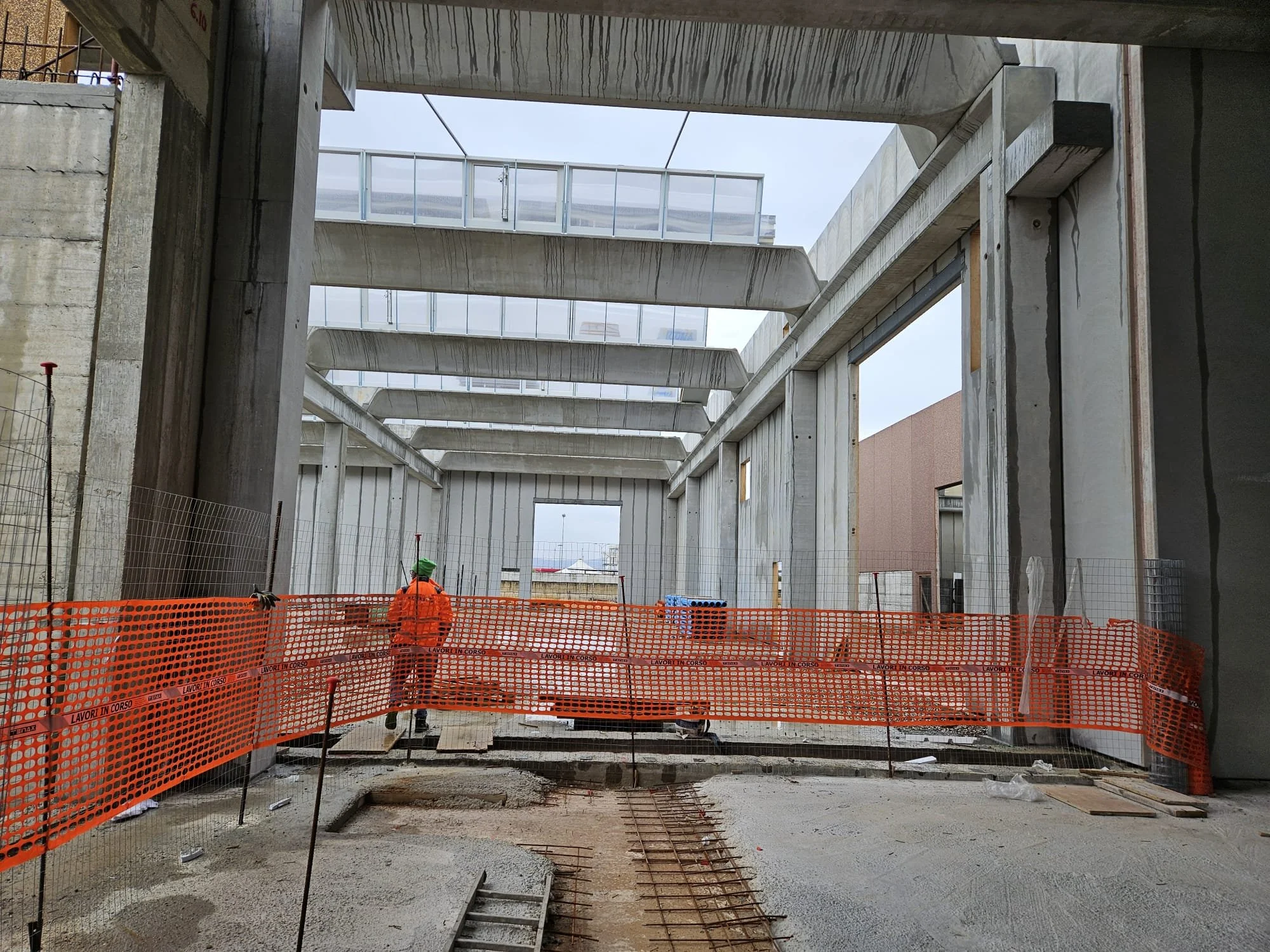 Cantiere edile con un lavoratore in gilet arancione e casco verde, recinzione di sicurezza arancione, struttura in cemento e tralicci in metallo.