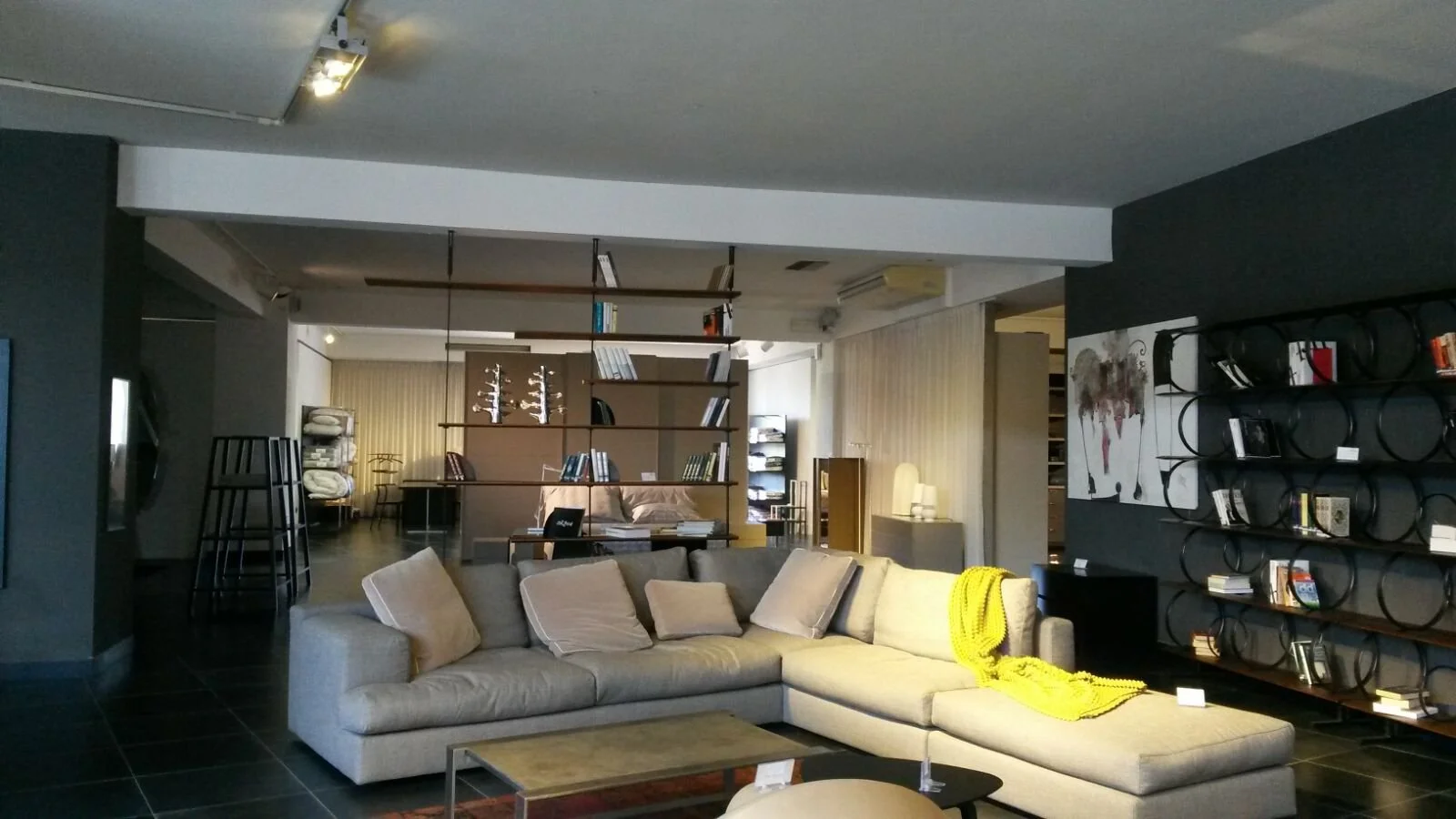 Soggiorno con divano beige, scaffale circolare nero con libri, pareti scure, lampade e decorazioni moderne.