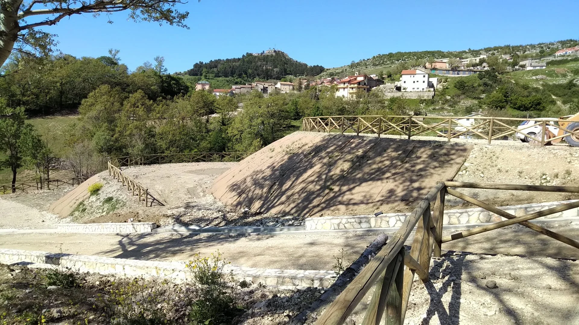 Paesaggio collinare con terrapieni in fase di modifica, recinzioni in legno e case sullo sfondo, sotto un cielo blu, in un'area rurale.