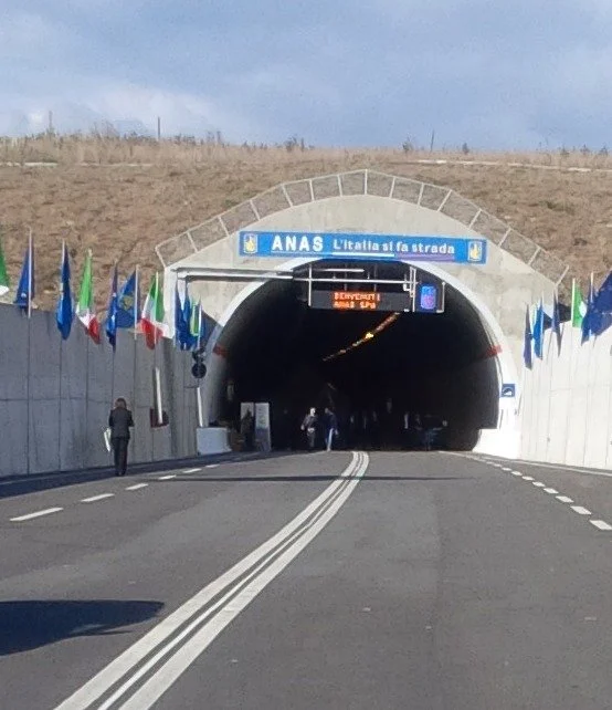 Ingresso di un tunnel autostradale con segnali di benvenuto e bandiere italiane e europee lungo i lati, ingresso della società ANAS, in Italia.