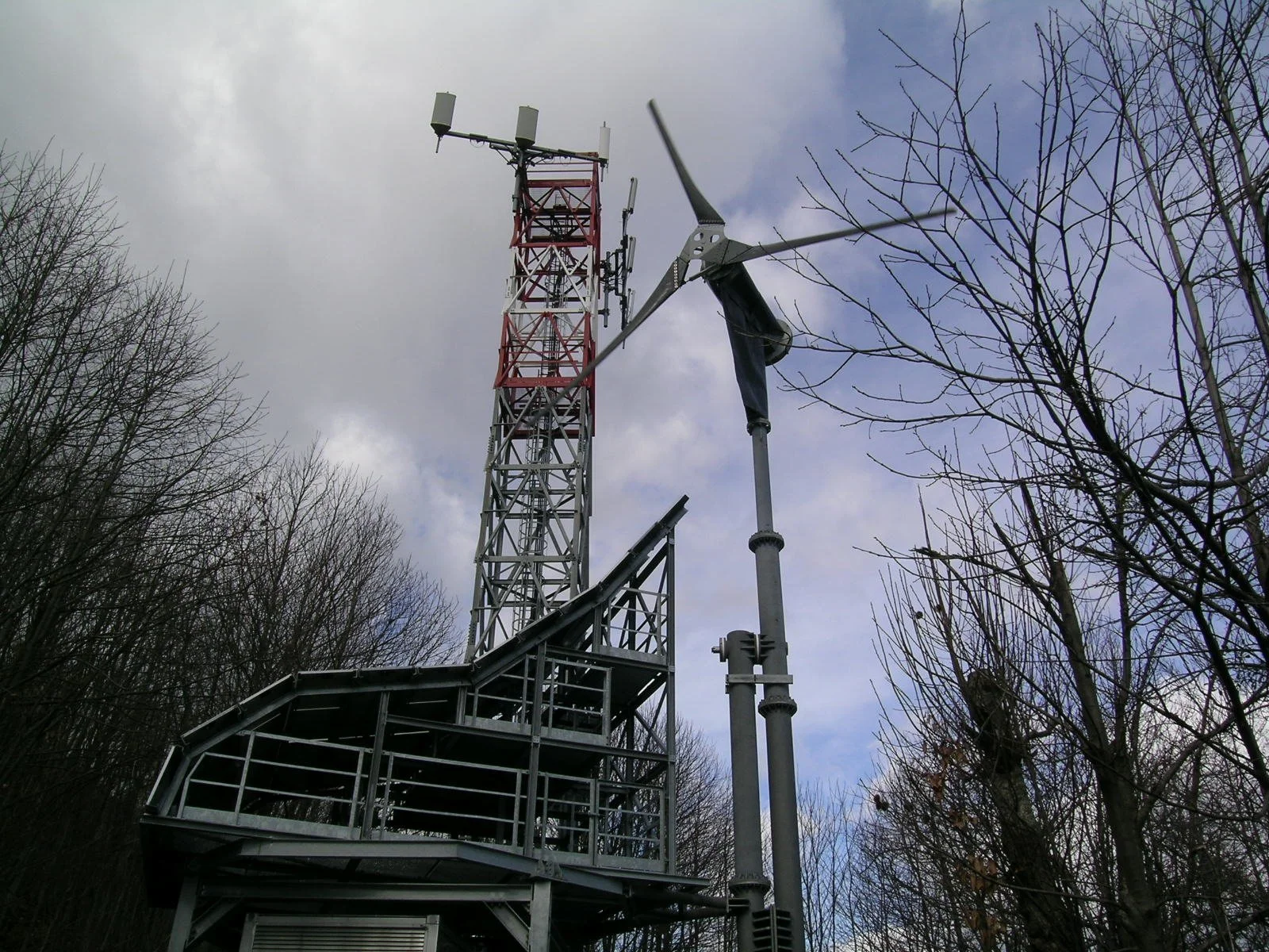 Torre di telecomunicazioni con pale eoliche, circondata da alberi senza foglie sotto un cielo nuvoloso.