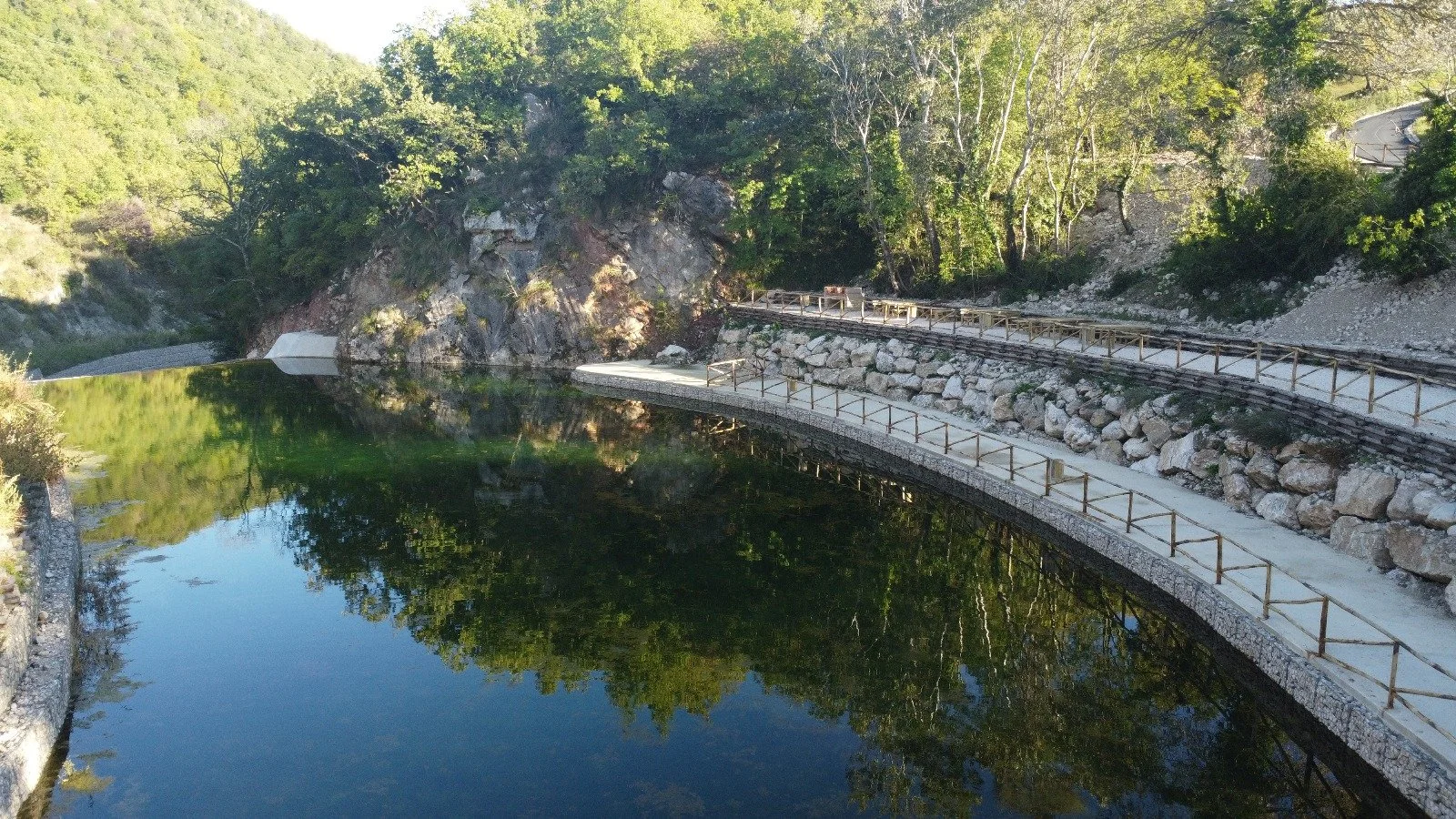 Sentiero accanto a un fiume con acque calme che riflettono gli alberi e le rocce circostanti, in un ambiente naturale con vegetazione lussureggiante.
