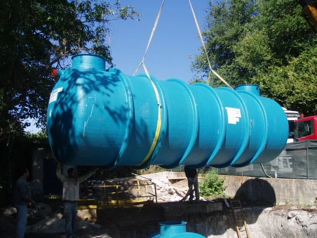 Un grande serbatoio blu viene sollevato da una gru mentre alcune persone assistono all'operazione in un'area di cantiere all'aperto.