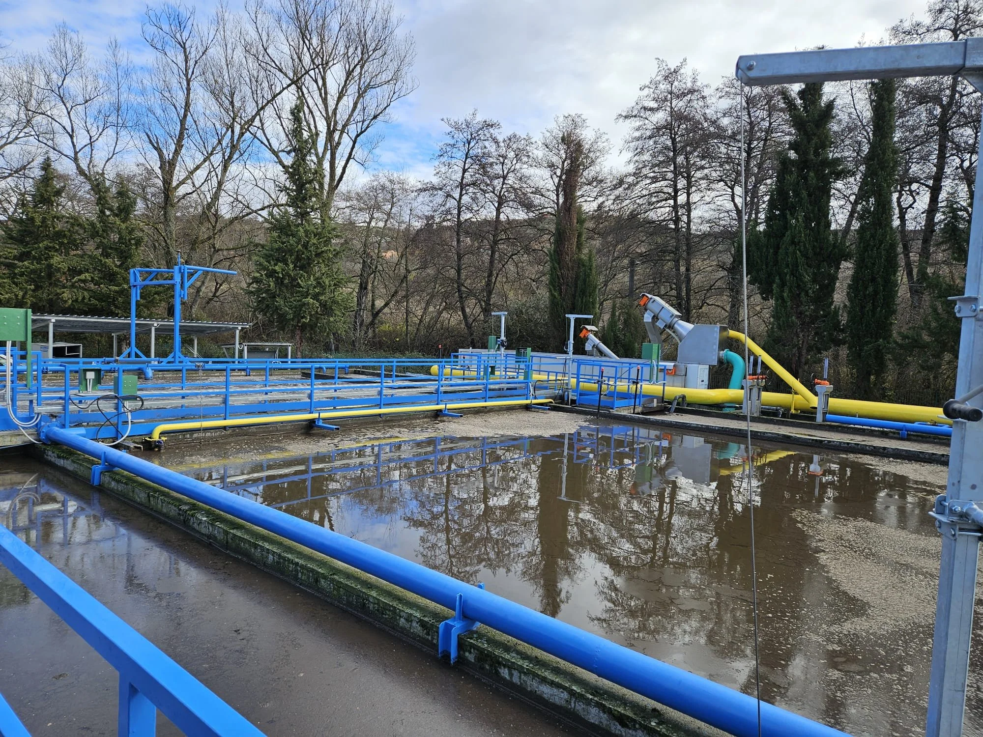 Impianto di trattamento delle acque reflue con tubature blu e getti di acqua a riposo, circondato da alberi spogli in un giorno nuvoloso.
