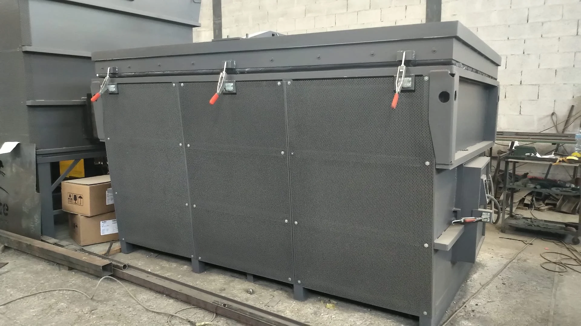 Macchinario industriale grigio con pannelli perforati, situato in un'officina con pareti in mattoni e scaffali con strumenti e materiali.