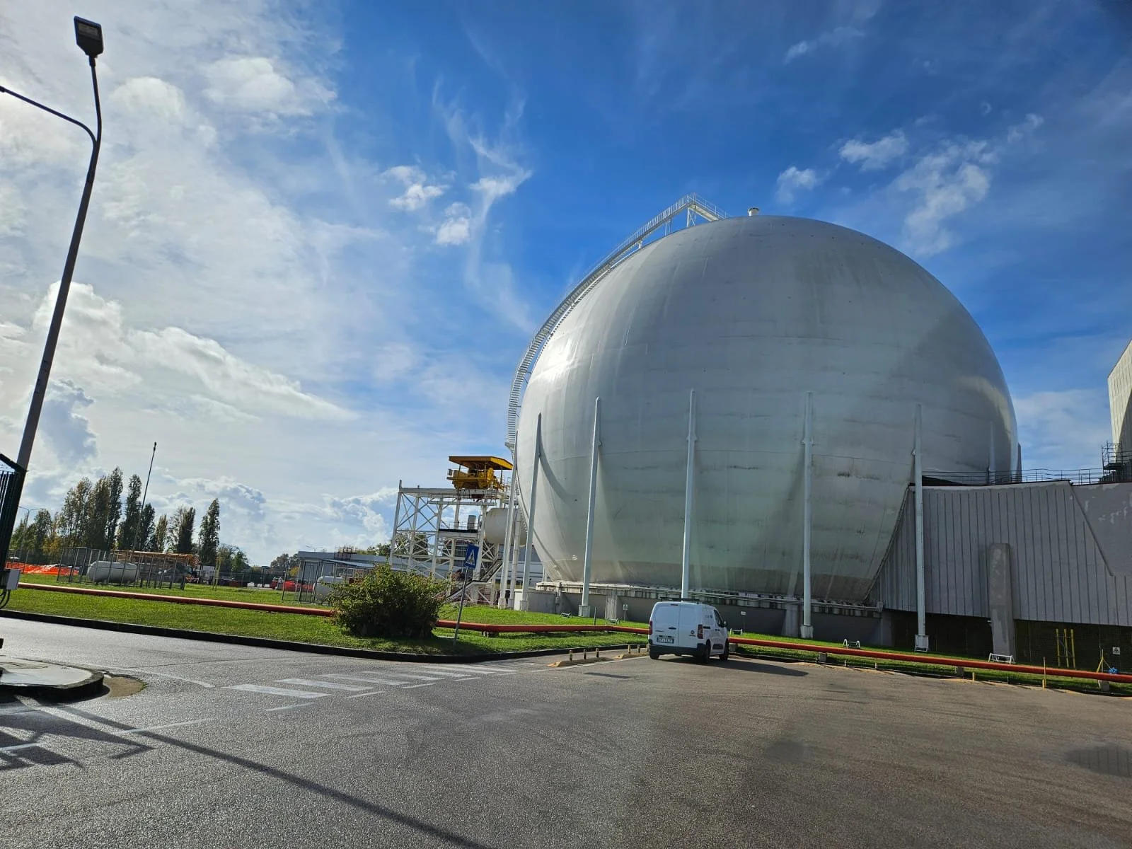 Grande serbatoio di forma sferica in un complesso industriale, sotto un cielo azzurro con nuvole sparse.