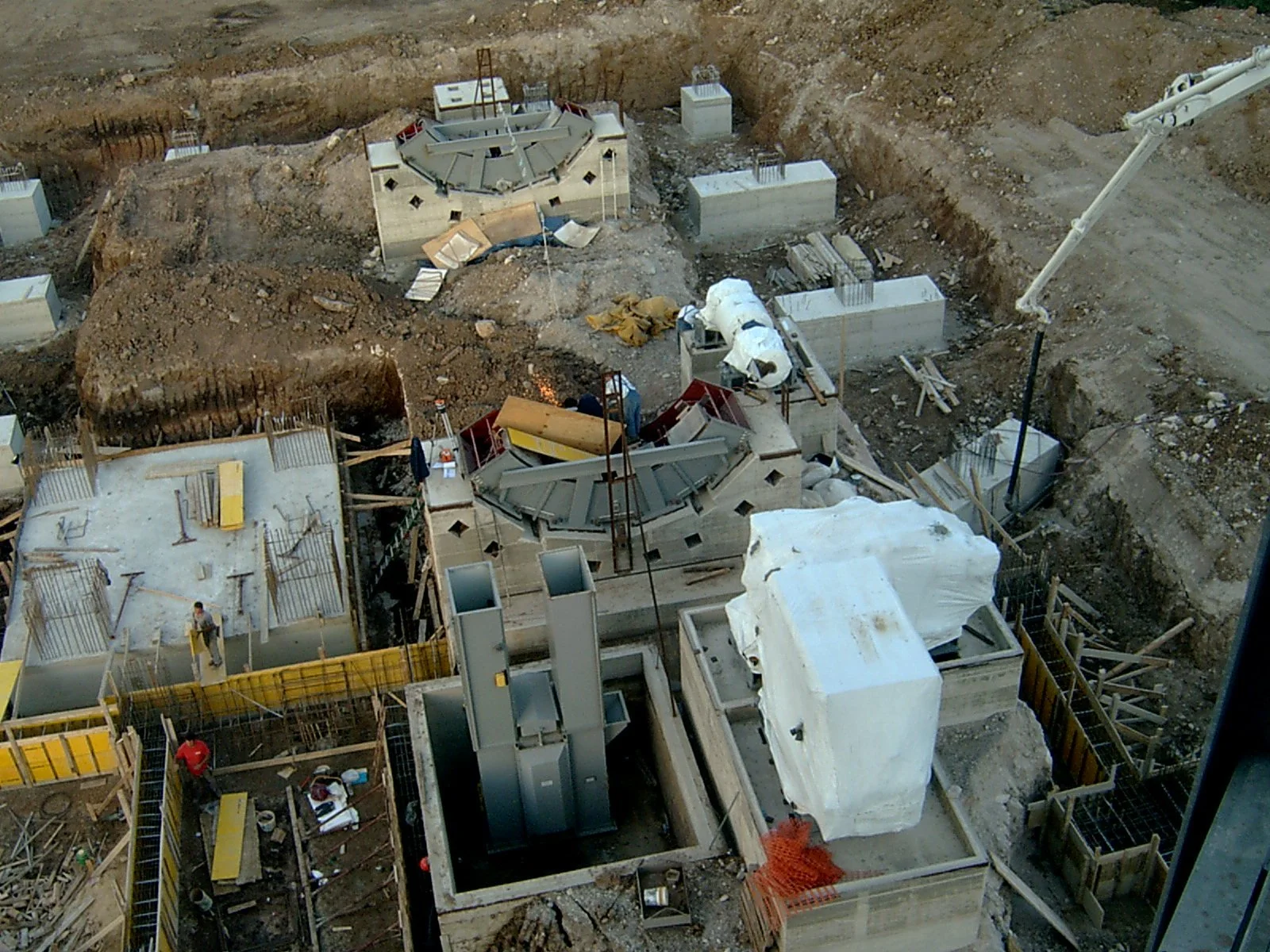 Immagine di un cantiere di costruzione con strutture in cemento, tubi e impalcature, mentre vari operai lavorano sul sito.