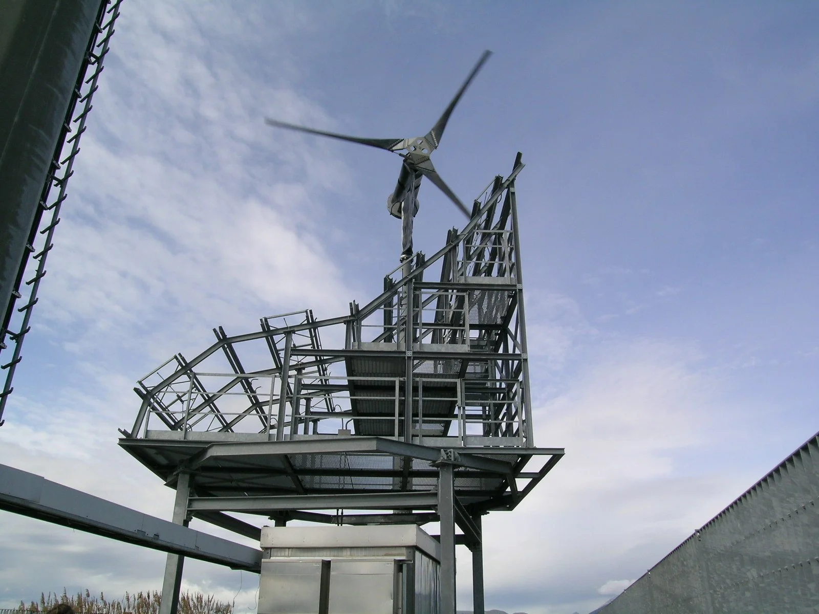 Pannello di controllo di una turbina eolica in metallo con pale al vento e cielo nuvoloso sullo sfondo.