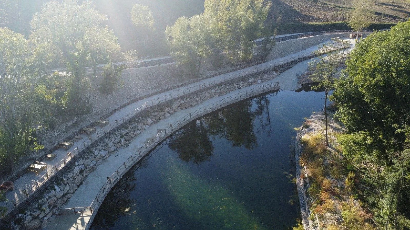 Vista aerea di un sentiero che corre lungo un fiume circondato da alberi verdi, con alcune panchine posizionate lungo il percorso.