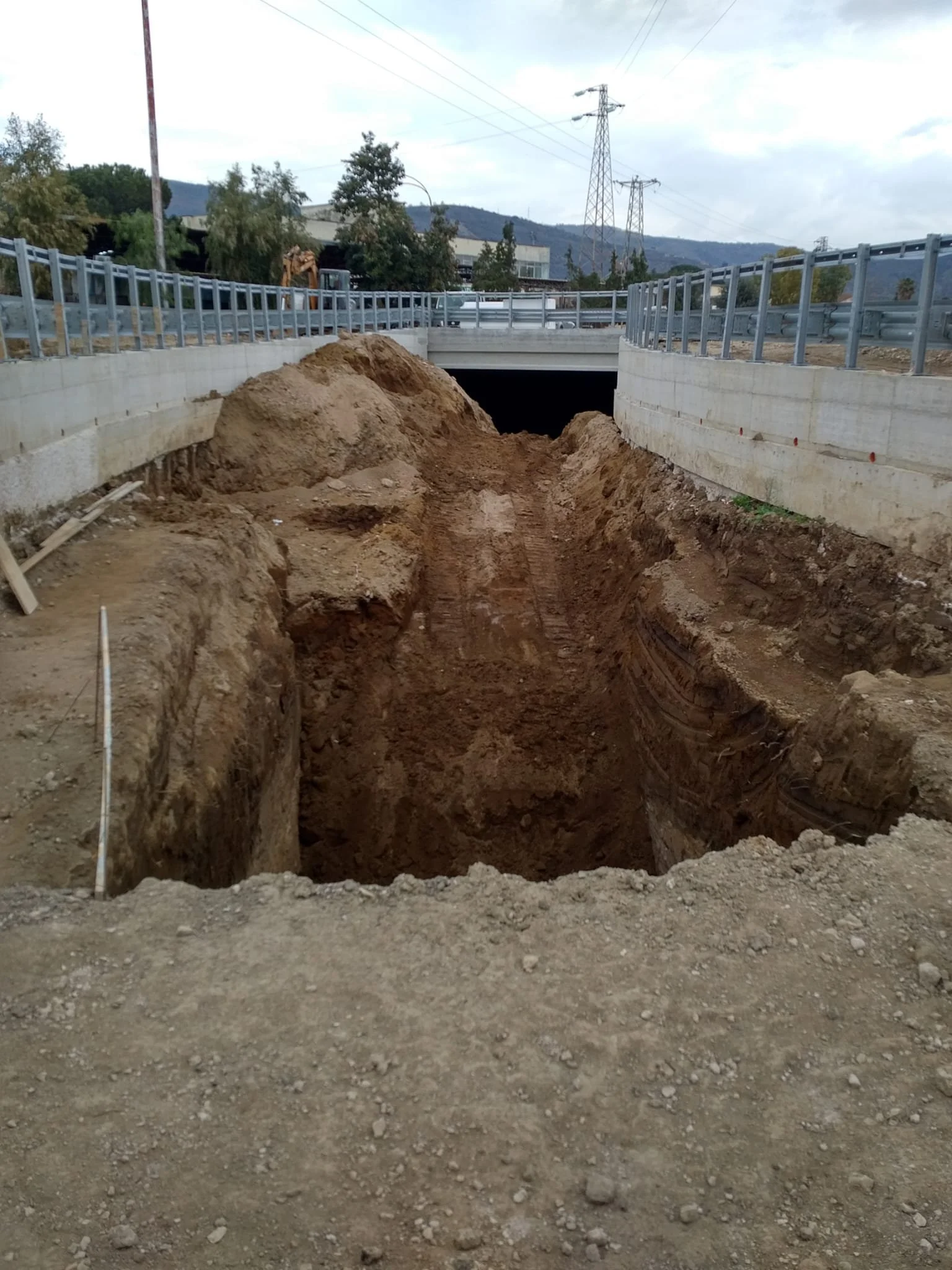 Cantiere stradale con scavi e terreno smosso sotto un ponte, con sfondo di alberi, montagne e linee elettriche.