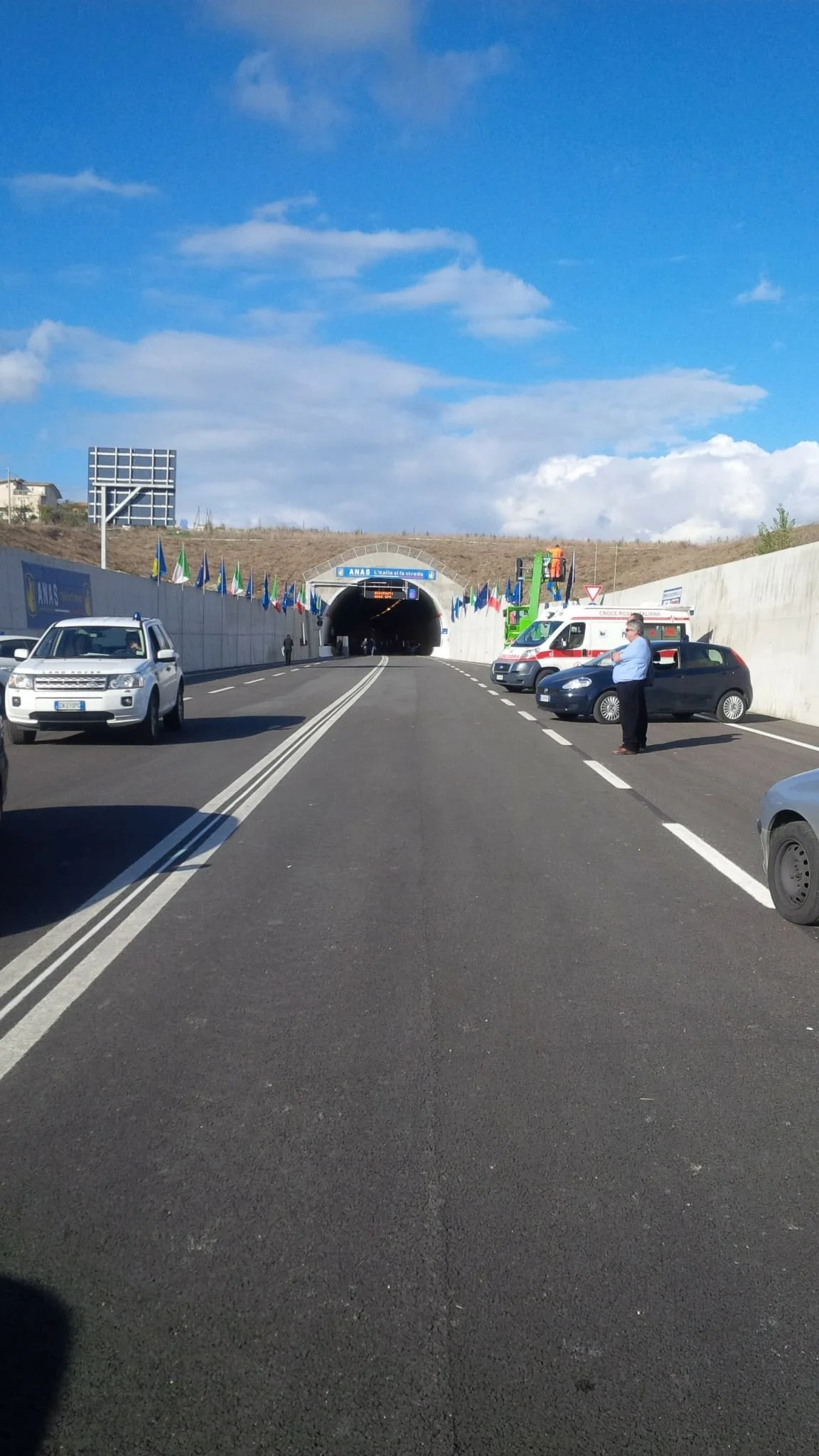 Ingresso di un tunnel autostradale con alcune macchine parcheggiate e alcune persone intorno, sotto un cielo nuvoloso.