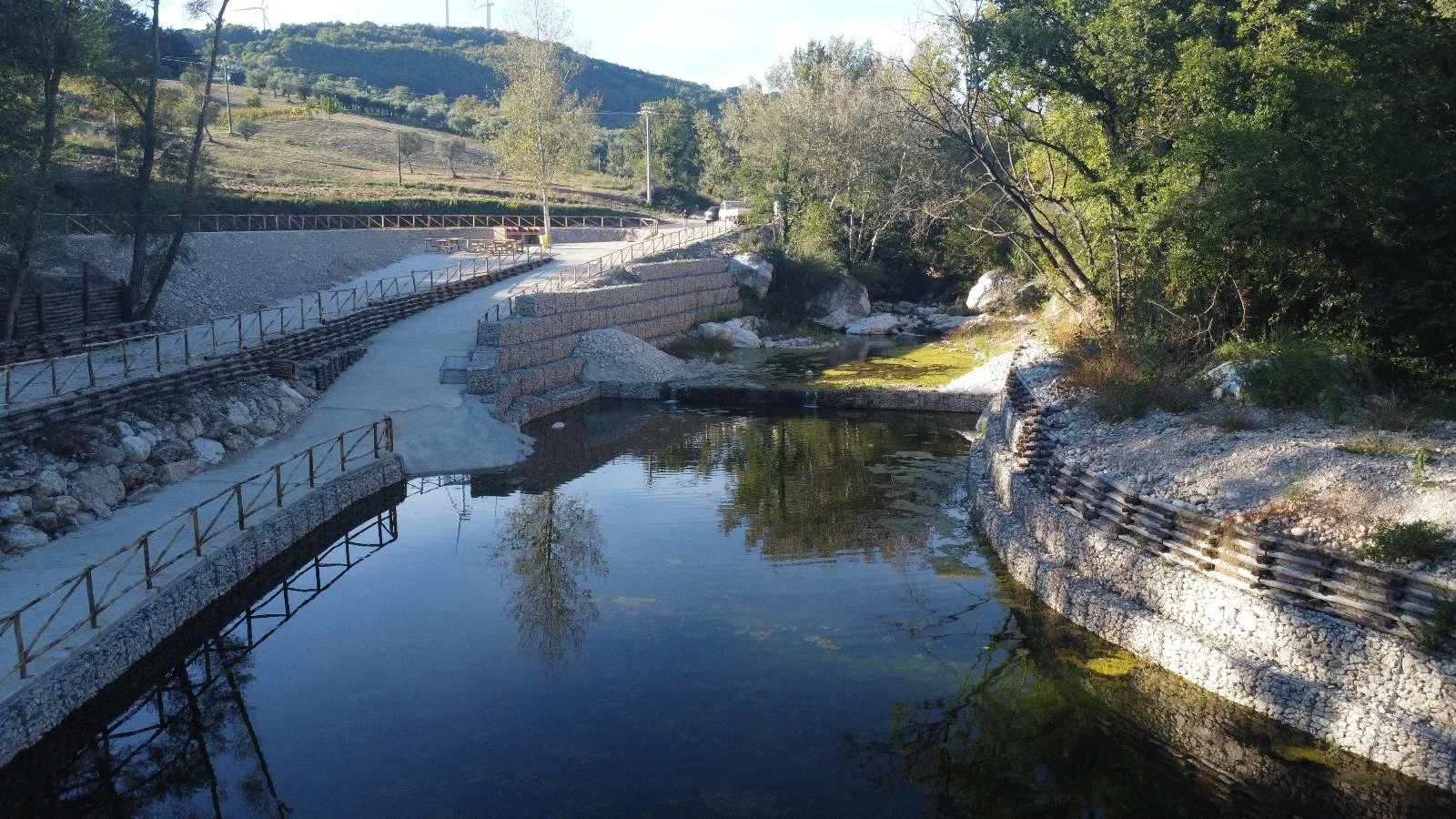 Una passeggiata lungo un corso d'acqua in un'area naturale, con recinzioni lungo il sentiero e alberi che circondano l'acqua, in un paesaggio collinare.