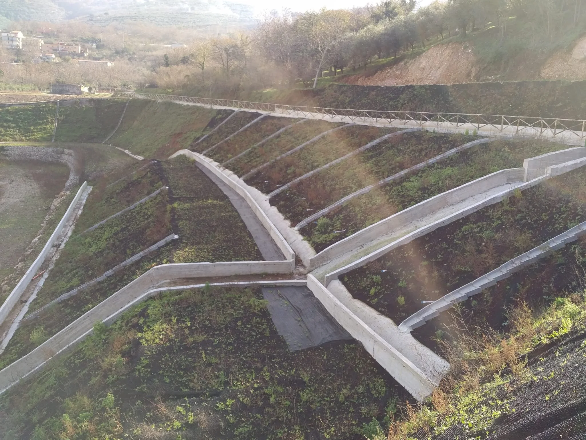 Terreno in pendenza suddiviso in sezioni con muri di contenimento e una passerella sopra. Vegetazione e terreni coltivabili sono visibili.