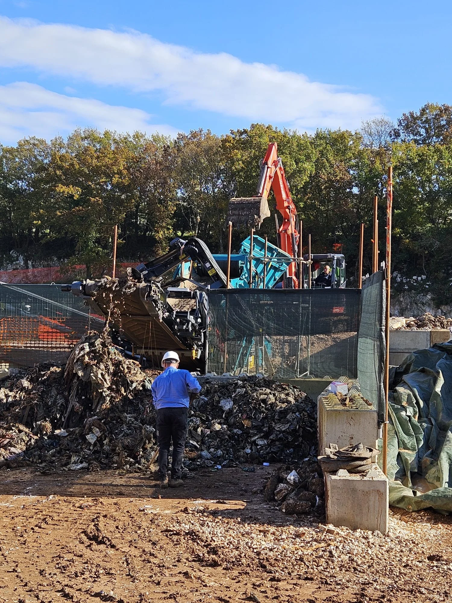Immagine di un sito di demolizione con un escavatore rosso e uno nero al lavoro, un uomo con casco e giacca blu di fronte a una pila di detriti, recinzione di sicurezza e fondo di alberi con cielo azzurro e nuvole.