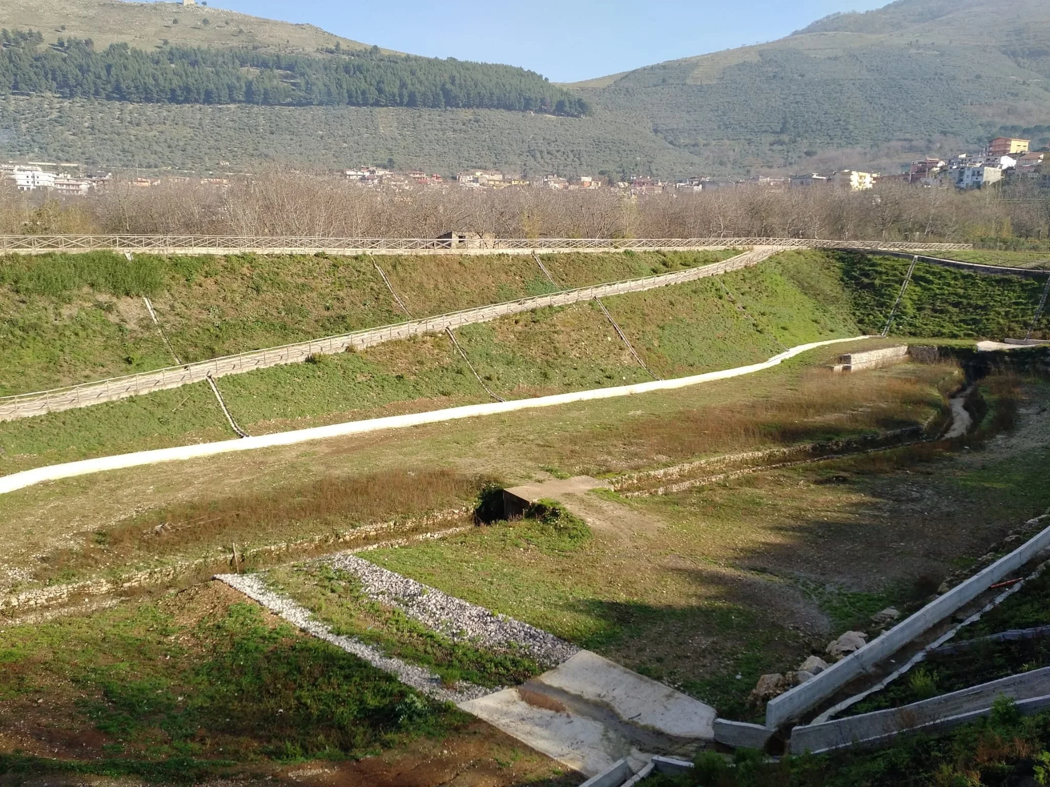 Colline con terreni e un sentiero sterrato, alcune strutture di cemento, sullo sfondo un paese e montagne verdeggianti.