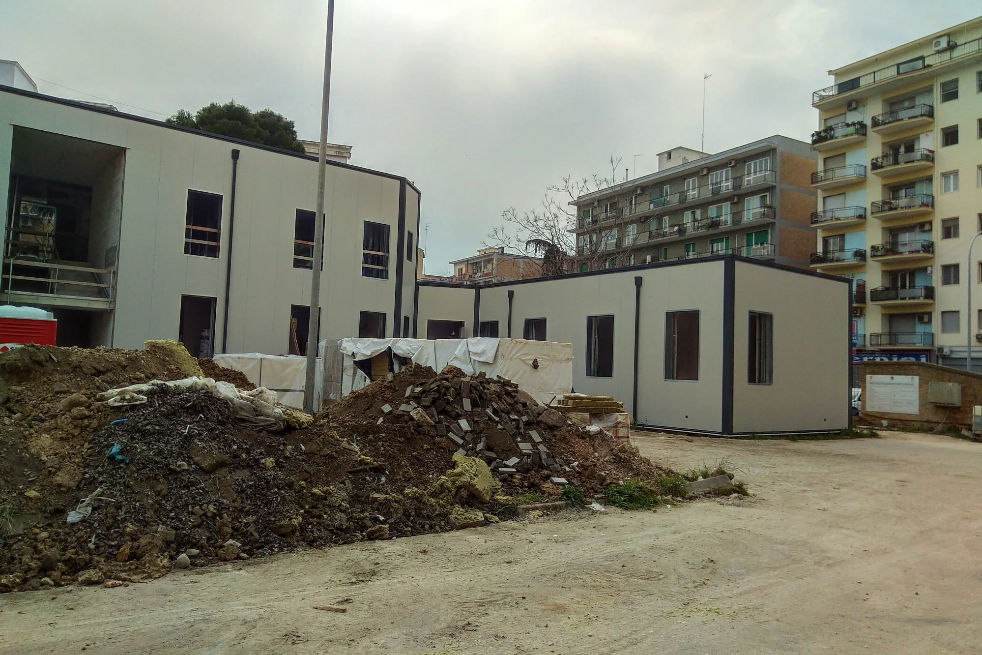 Cantiere edile con terreno di terra e pile di detriti, edifici residenziali intorno