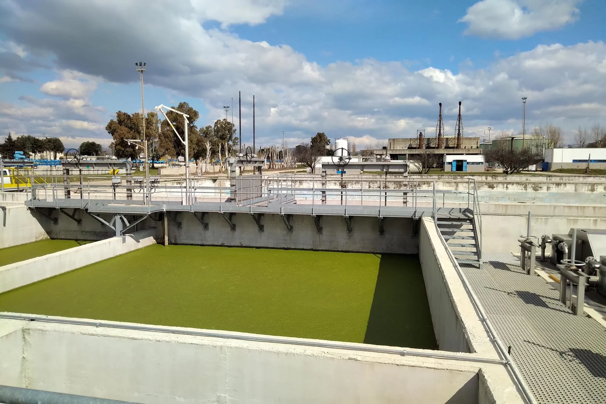 Stamme di depurazione dell'acqua con superfici di colore verde e strutture metalliche di supporto, sotto un cielo parzialmente nuvoloso.