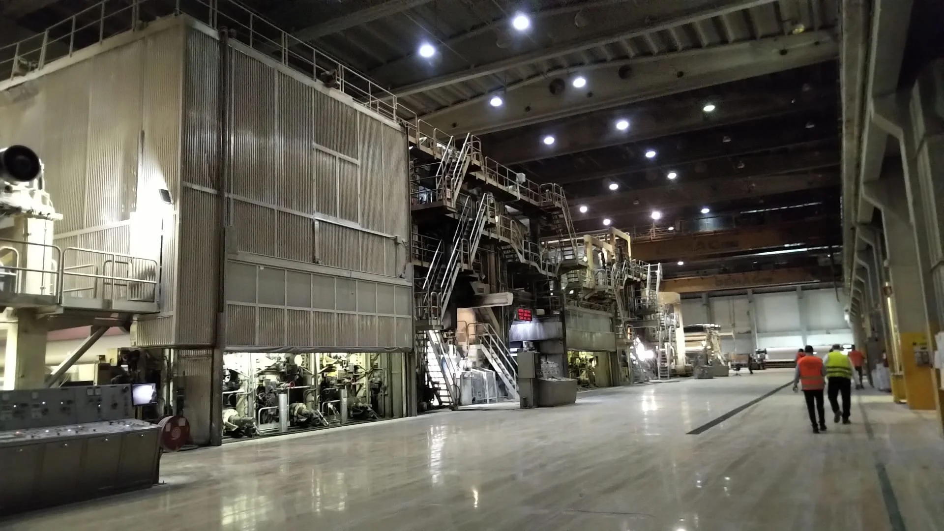 Interno di una centrale nucleare con grandi apparecchiature, passerelle e luci sul soffitto, con alcune persone in cammino.