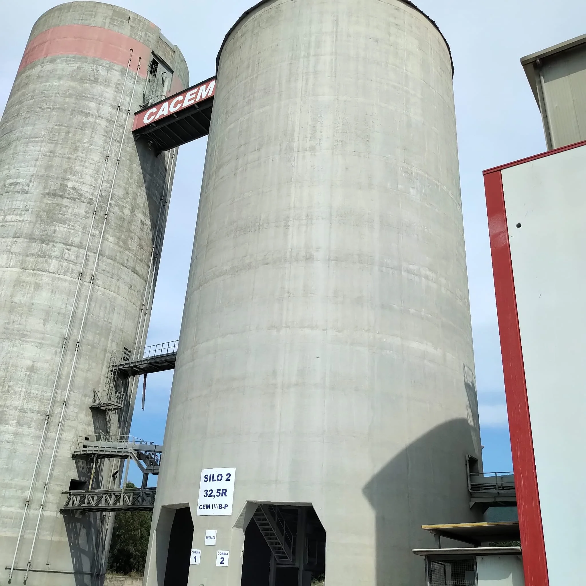 Immagine di due silos di cemento industriali, uno dei quali ha un cartellino che indica 'SILO 2 32,5 R CEM IV/B-P'. Sul silo c'è un'insegna con la scritta 'CACEM'.