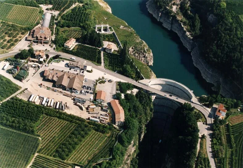 Vista aerea di una diga con un bacino idrico, una strada che attraversa la diga, edifici e aree di coltivazione circostanti, e un canyon naturale con acque che scorrono attraverso le rocce.