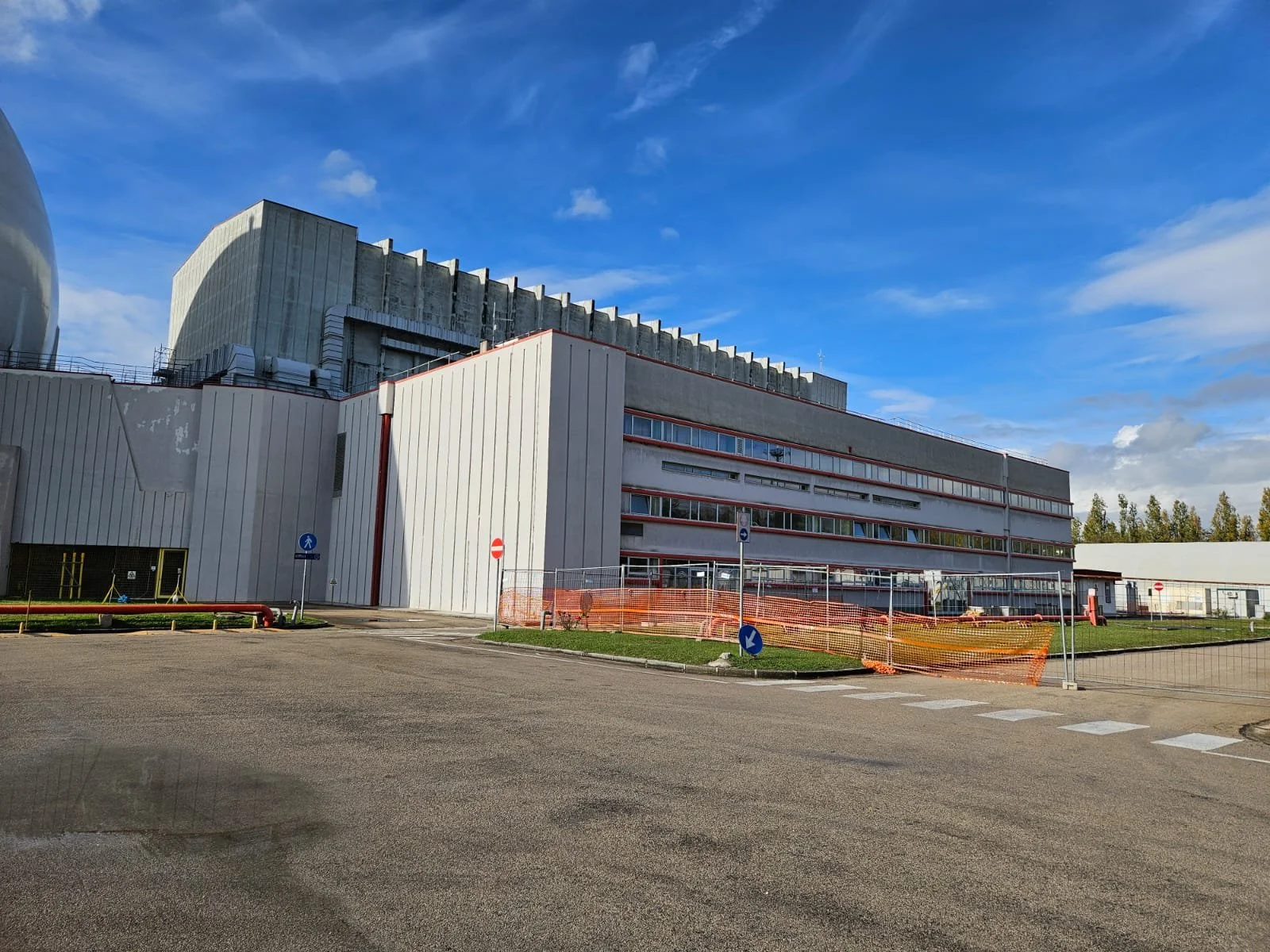 Edificio industrial o di ricerca con strutture moderne, cielo blu, pendenza di sicurezza arancione e cartelli stradali.