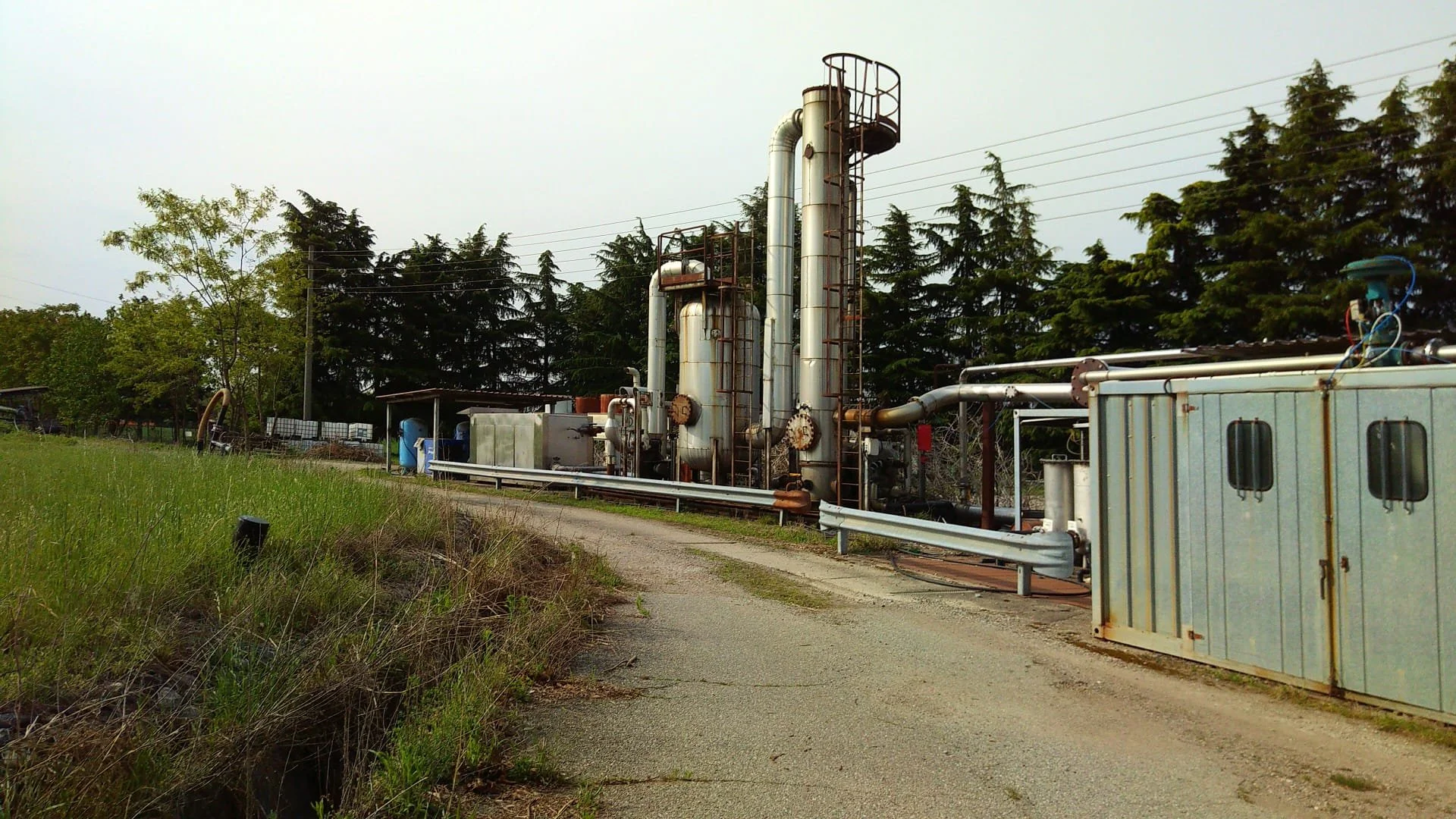 Impianto industriale con grandi tubi metallici e strutture in ferro, circondato da alberi e un sentiero in primo piano.
