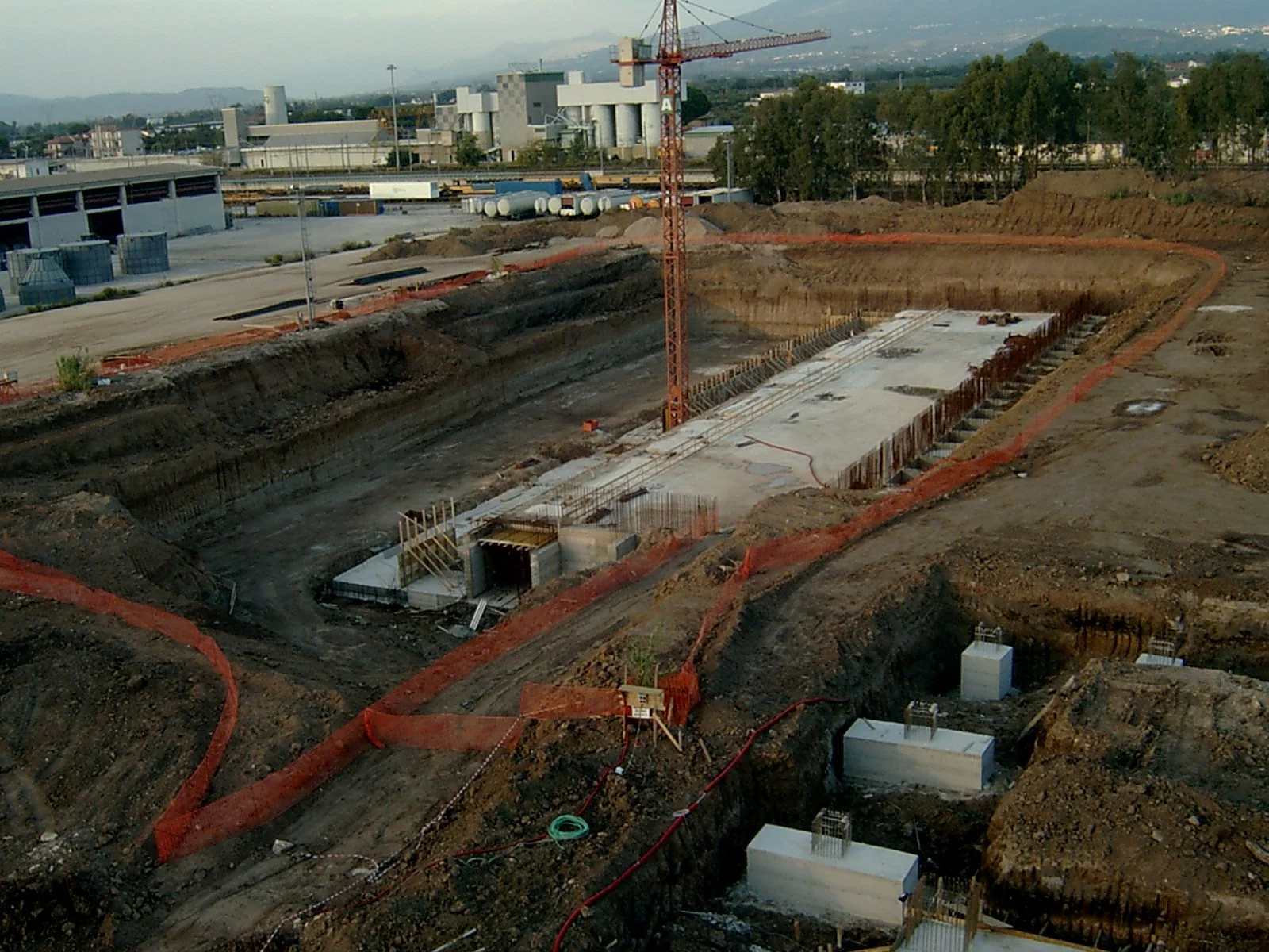 Cantiere edile con scavi profondi, fondamenta in costruzione, presenza di una gru rossa e recinzioni di sicurezza arancioni.