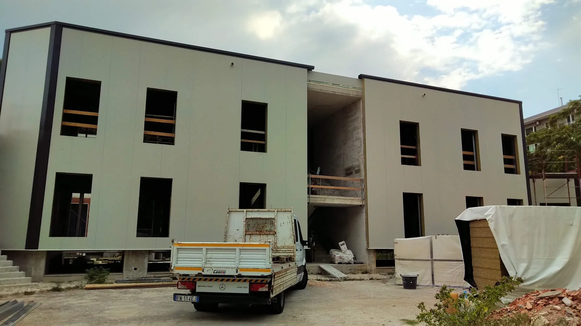 Edificio in costruzione con pareti bianche e apertura centrale, sviluppo a due piani, con camion e materiali di costruzione davanti.