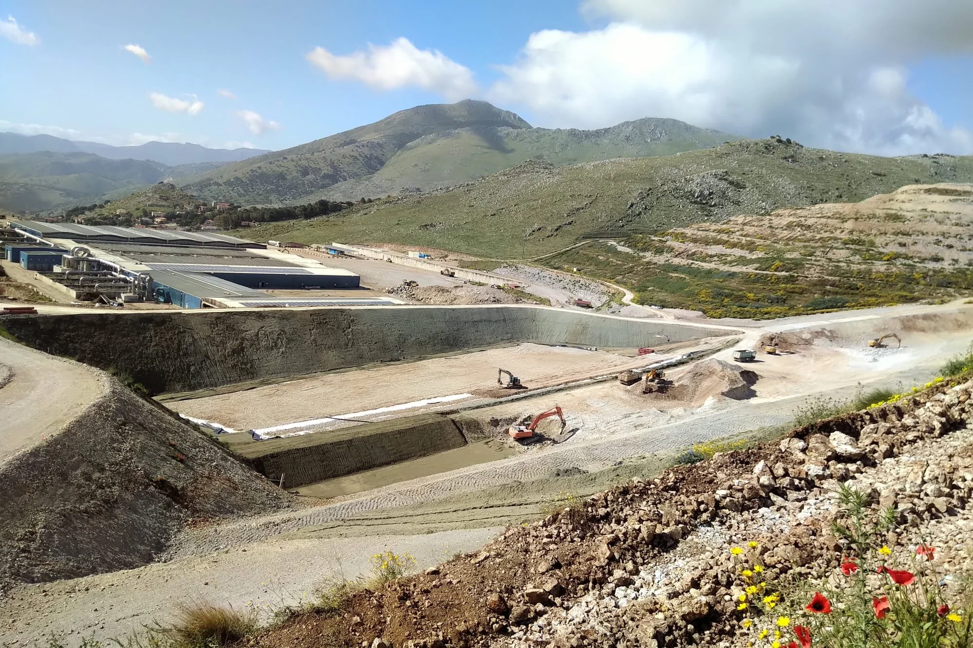 Immagine di un cantiere di costruzione in una valle montuosa con terreno in fase di scavo e alcune macchine da lavoro, sullo sfondo colline verdi e monti sotto un cielo parzialmente nuvoloso.