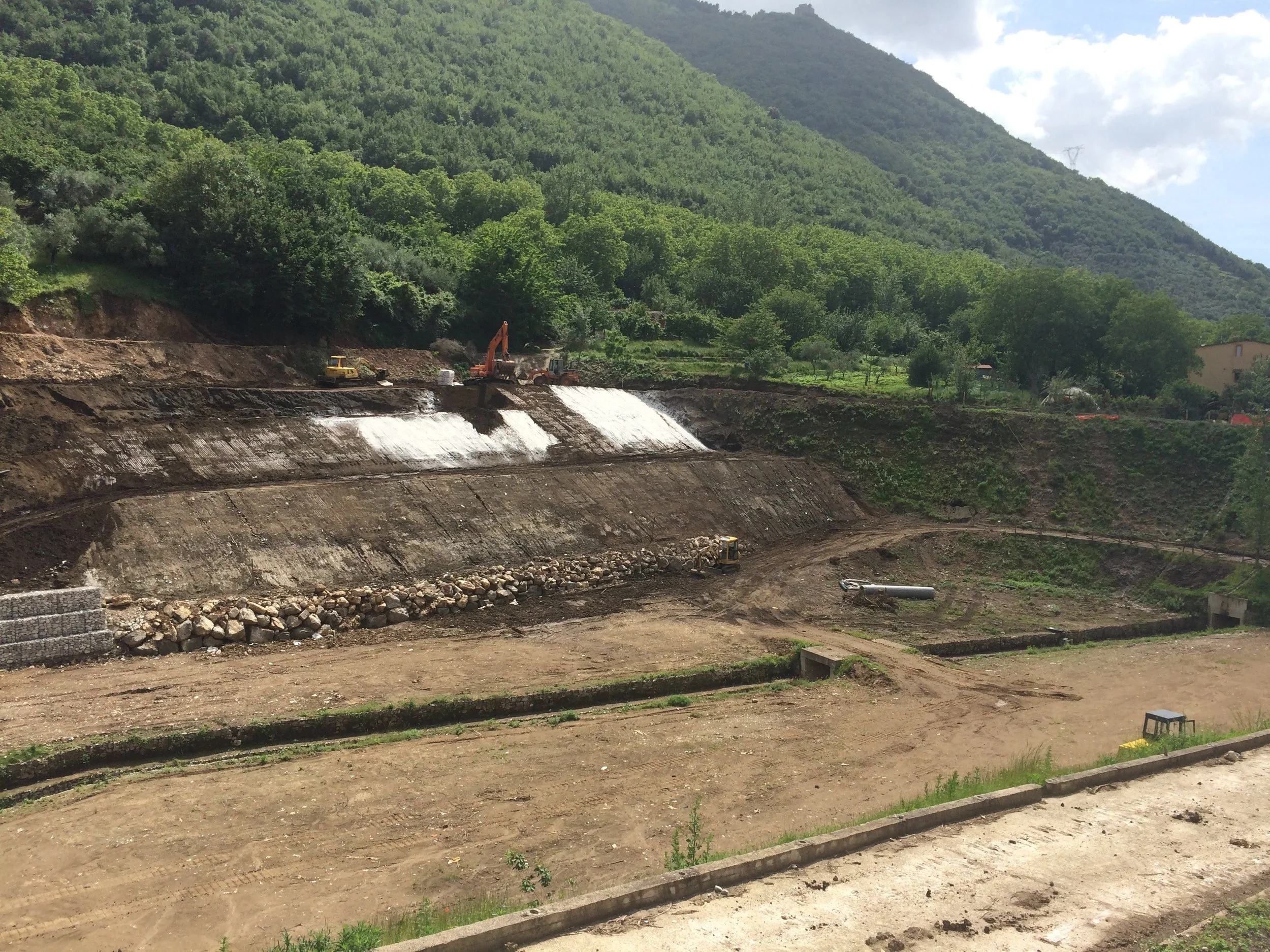 Lavori in corso su una collina con attrezzature da costruzione, scavi e terre movimentate, con un paesaggio montano verde sullo sfondo.
