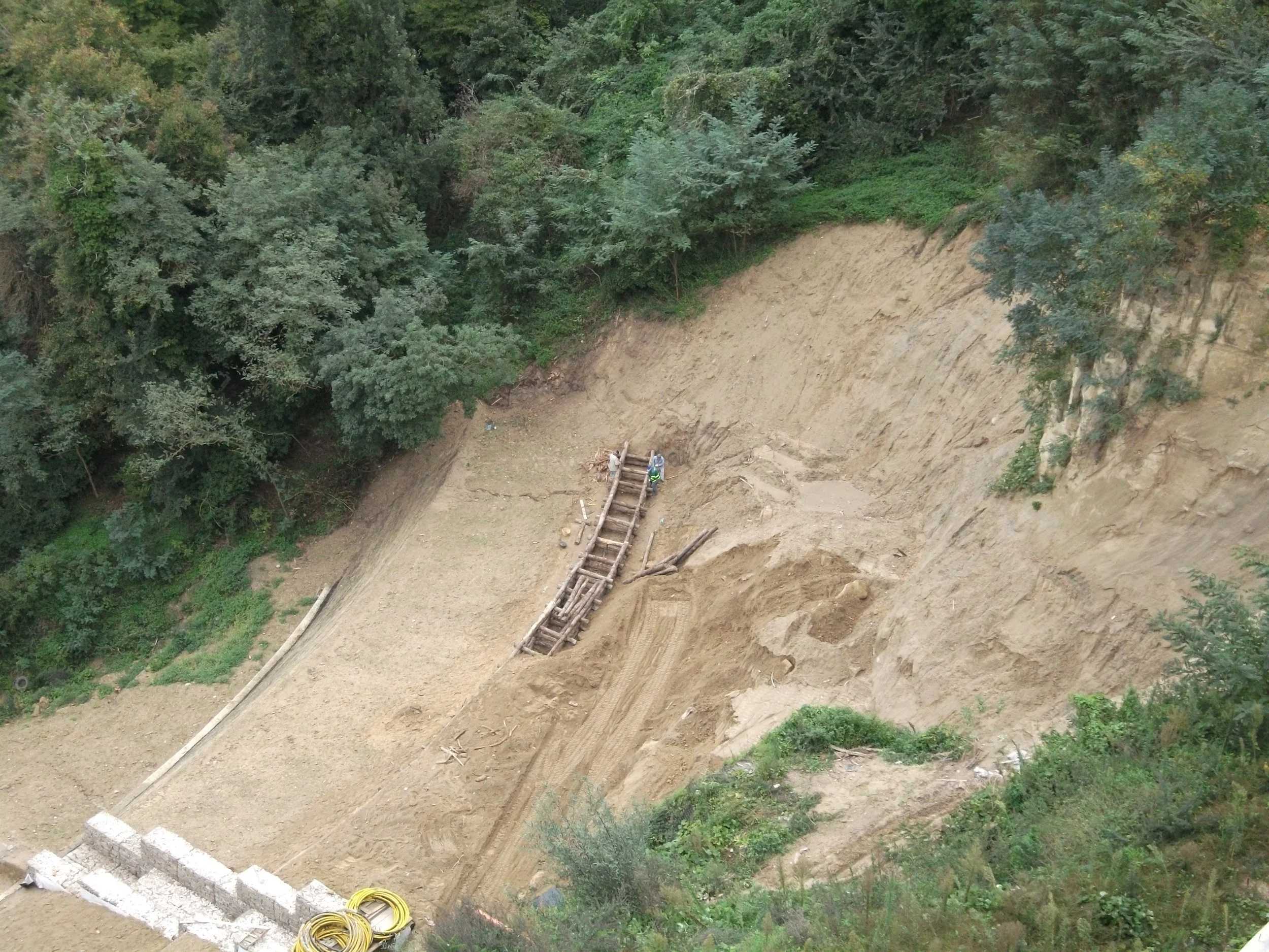 Scavi in corso su una collina con escavatore e due operai, con vegetazione verde in fondo.