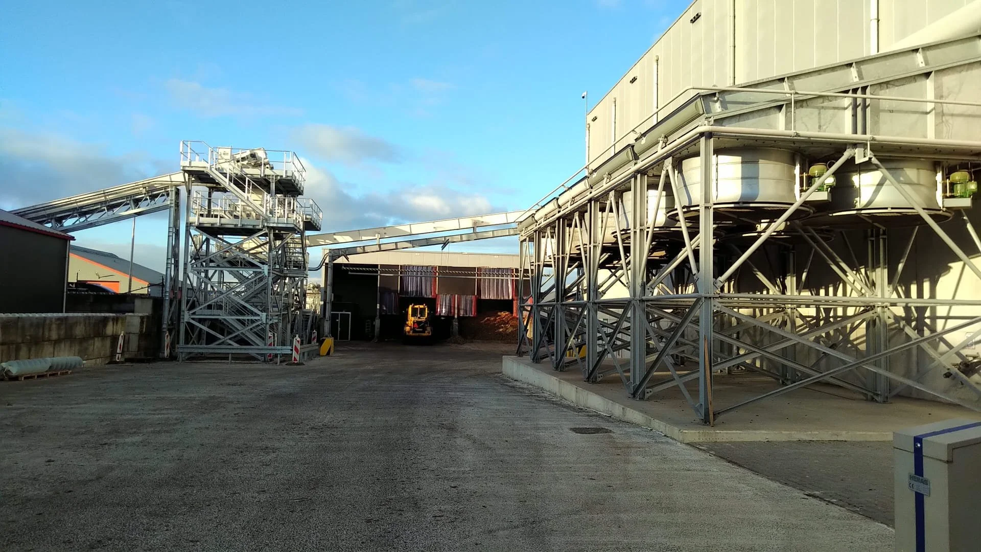 Impianto industriale con strutture metalliche e tubature, strada asfaltata e edificio sullo sfondo, sotto un cielo con nuvole.