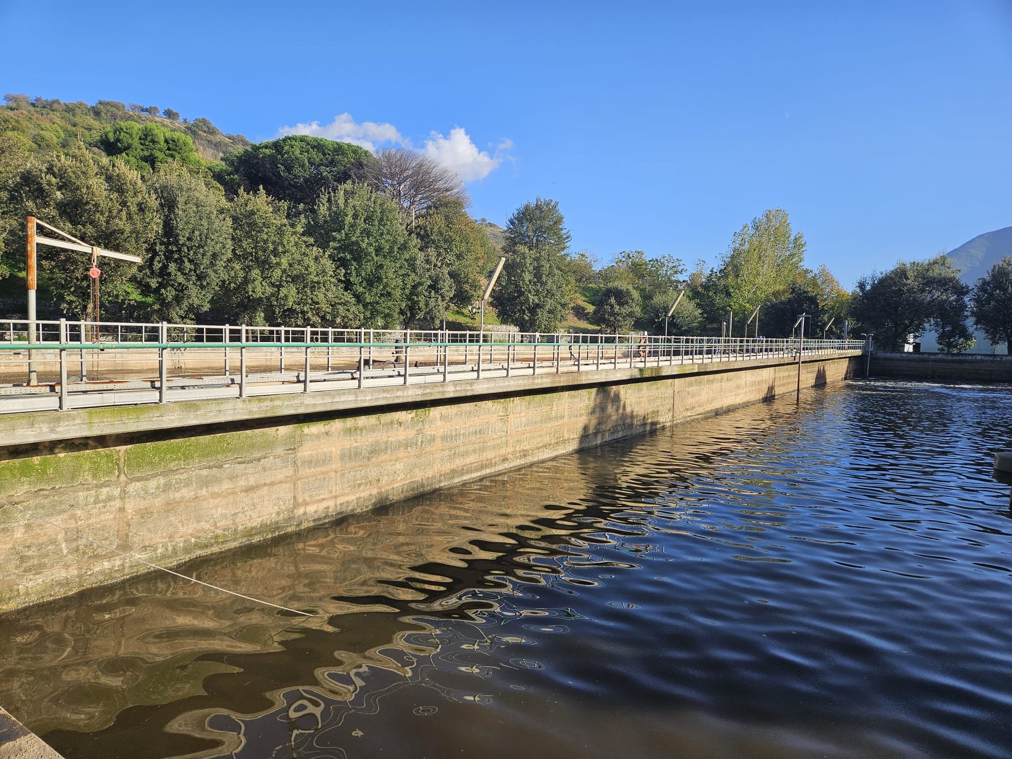 Lungo un muro di cemento che separa un'acqua tranquilla, si vedono alberi verdi e colline sullo sfondo sotto un cielo blu con alcune nuvole.