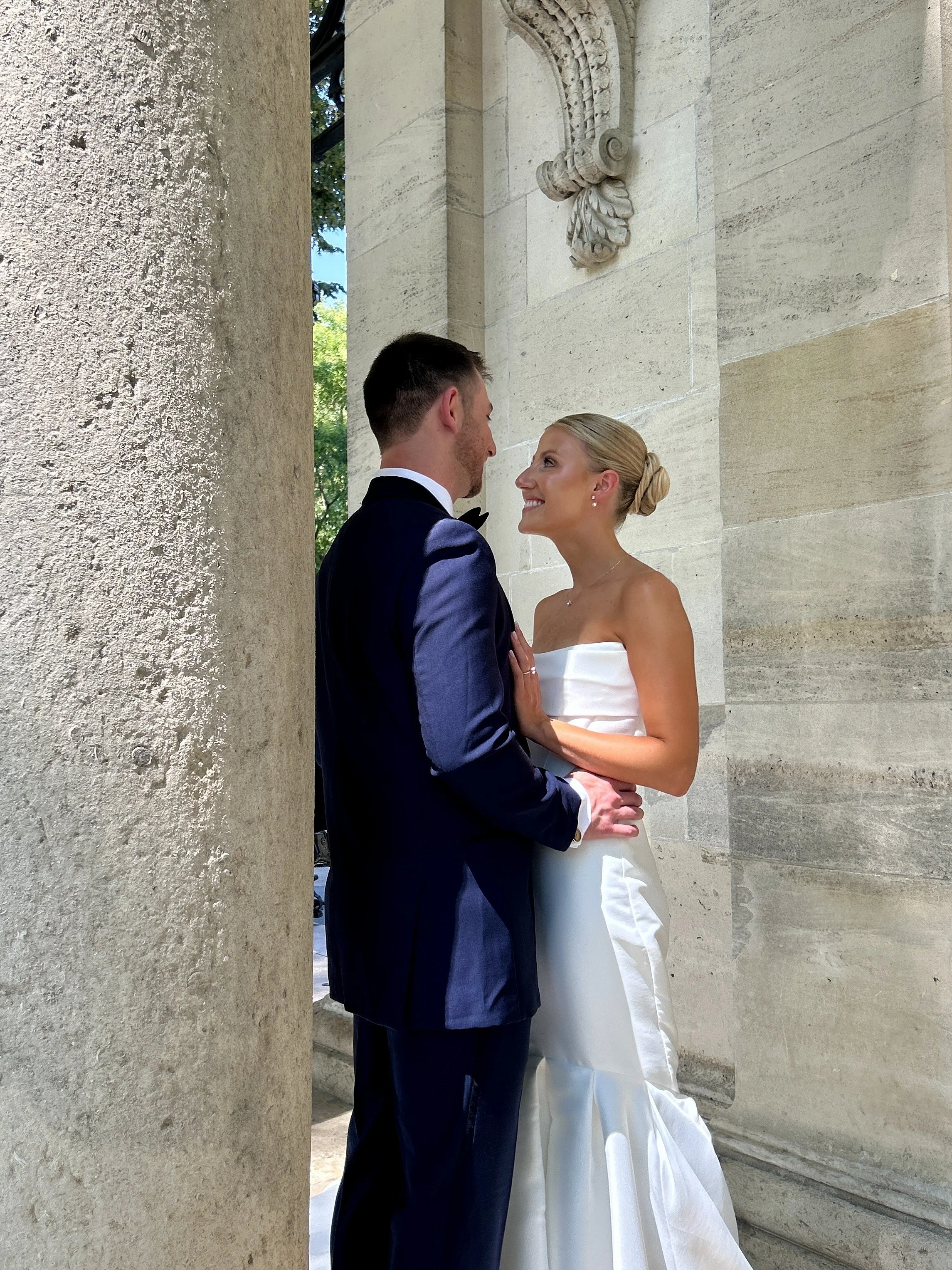 A bride and groom on their wedding day, standing close and smiling at each other near a stone column and wall with decorative carvings.