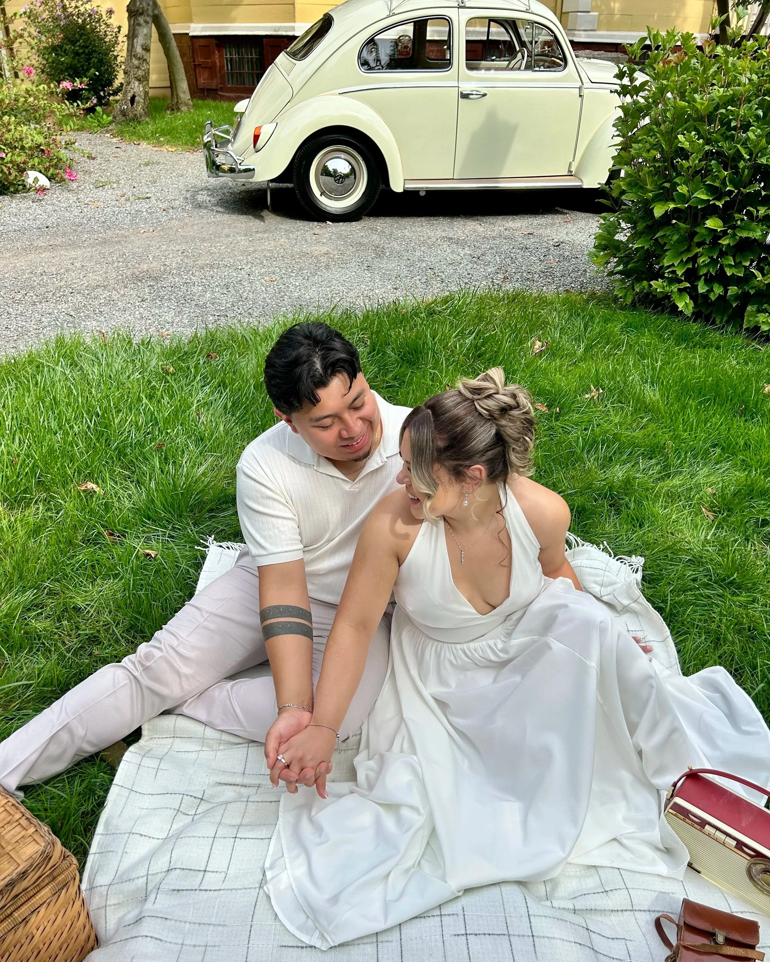 A couple sitting on a blanket outdoors, smiling and holding hands, with a vintage white car and greenery in the background.