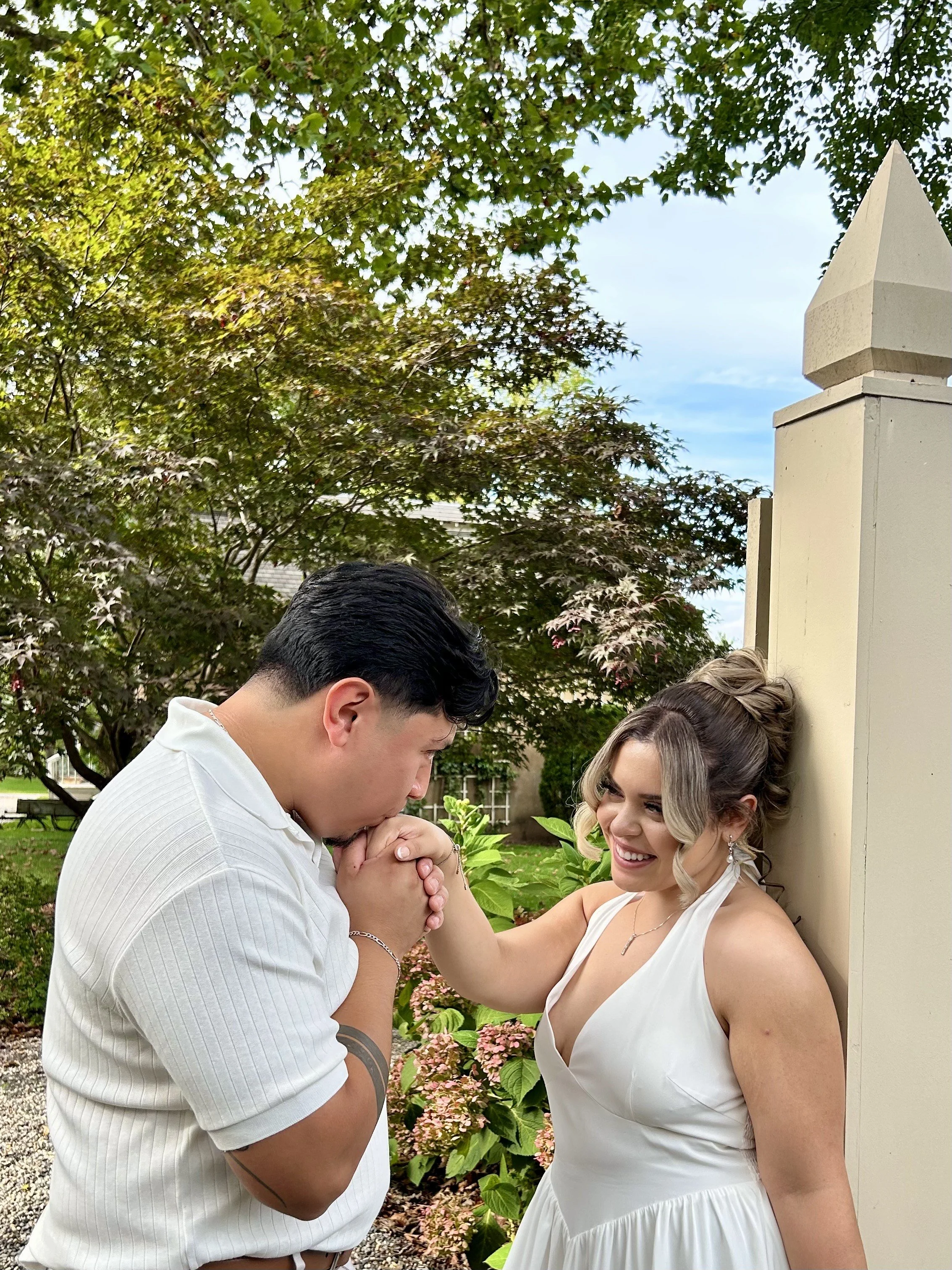 A man and woman share a moment outside, with the man holding the woman's hand and leaning in as if to kiss it. The woman is smiling and leaning against a beige wall, wearing a white dress with blonde hair styled in curls.