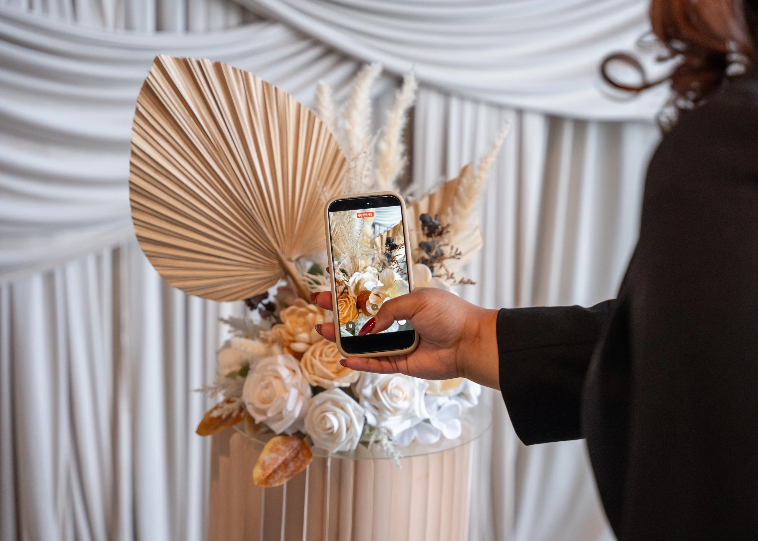 Person taking a photo of a floral arrangement with a smartphone. The arrangement includes white roses, dried leaves, and pampas grass, set against a draped fabric backdrop.