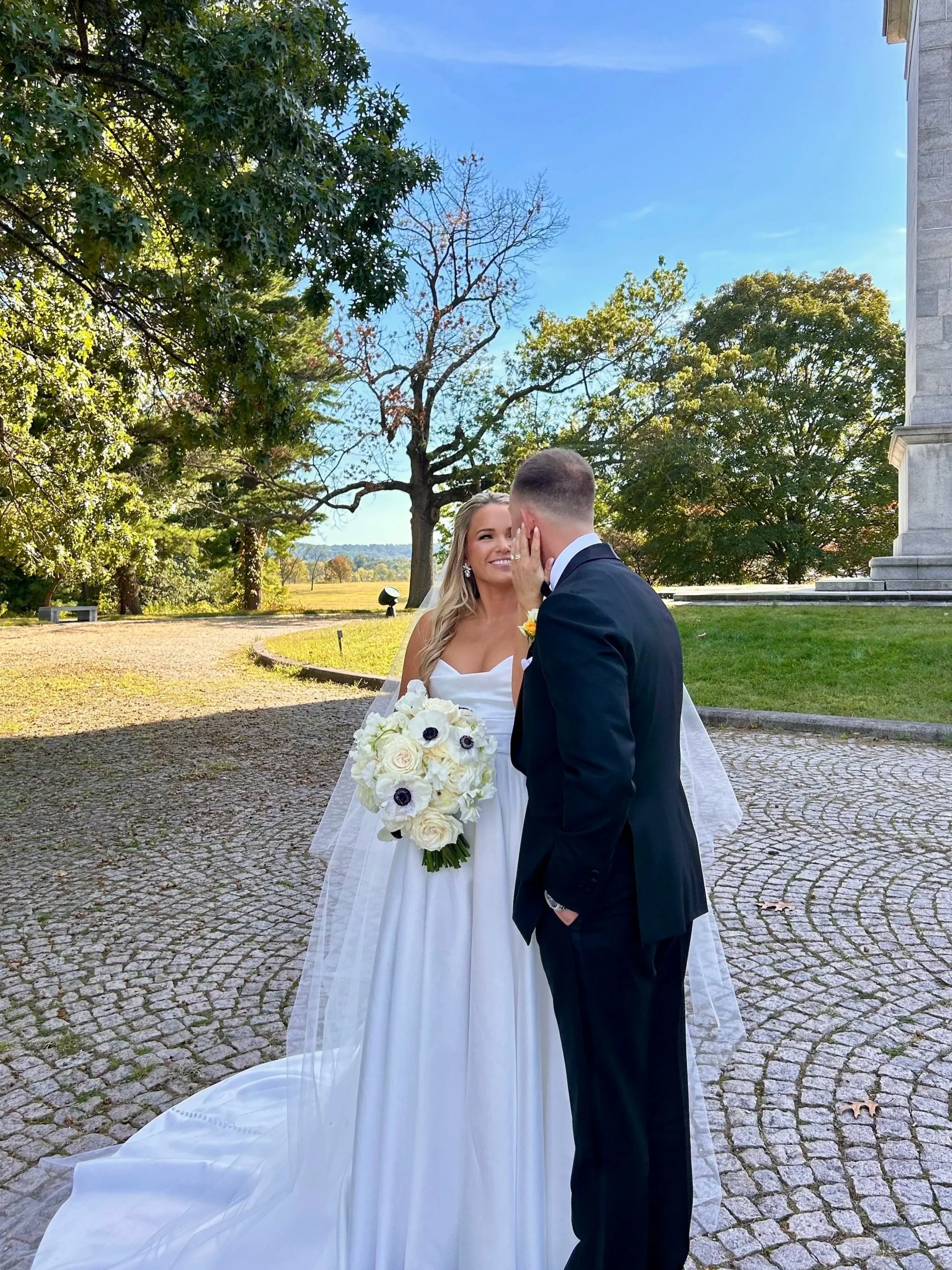 A bride and groom share a romantic moment outdoors on a sunny day, with the bride smiling and holding a bouquet of white flowers, and the groom gently touching her face.