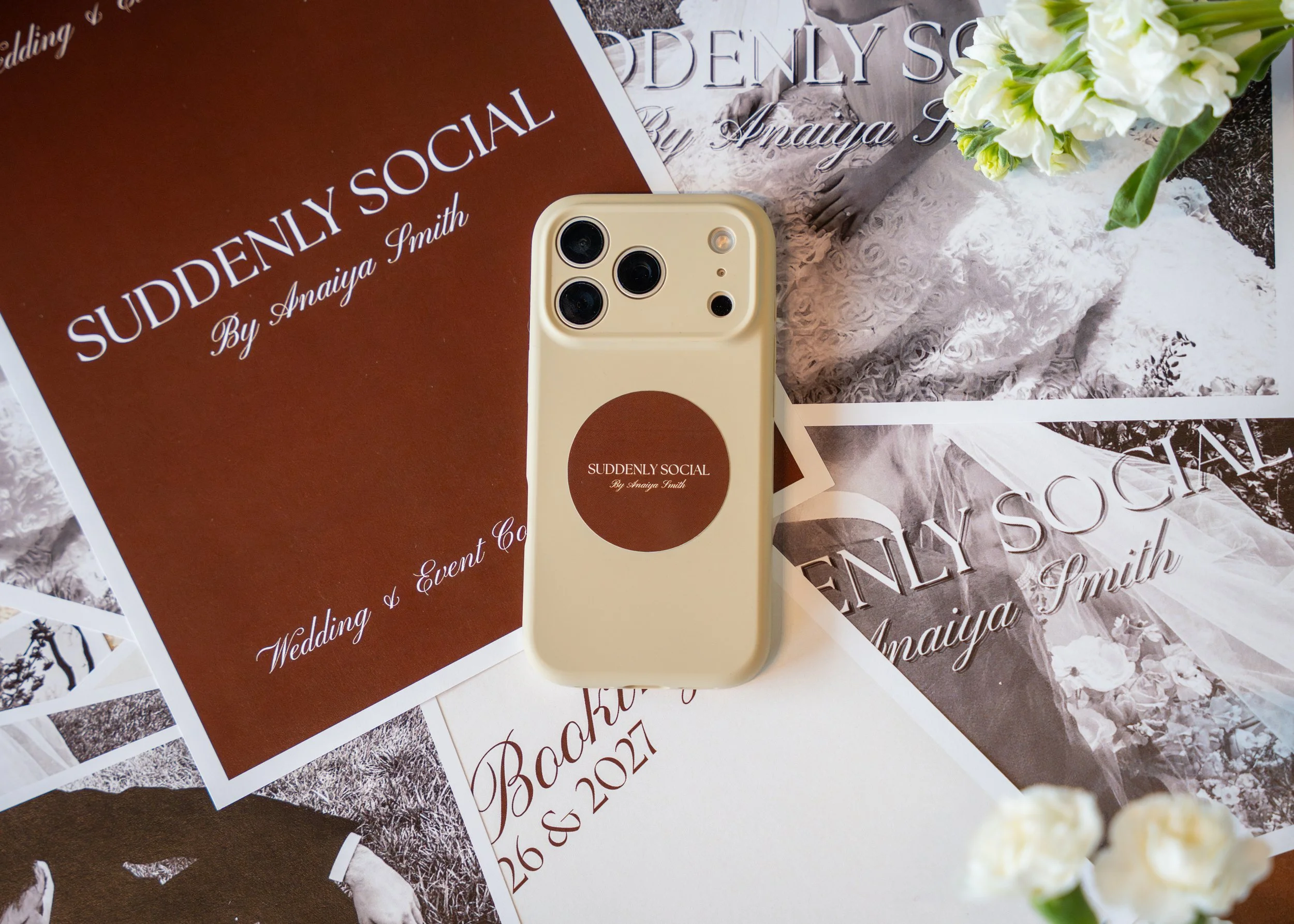 A beige iPhone with a brown circular sticker on the back that says 'Suddenly Social by Anaiya Smith' is placed on top of various wedding-themed papers and photographs, including one with the same title and subtitle, a wedding announcement, and a black-and-white wedding photo with a bouquet of white flowers nearby.