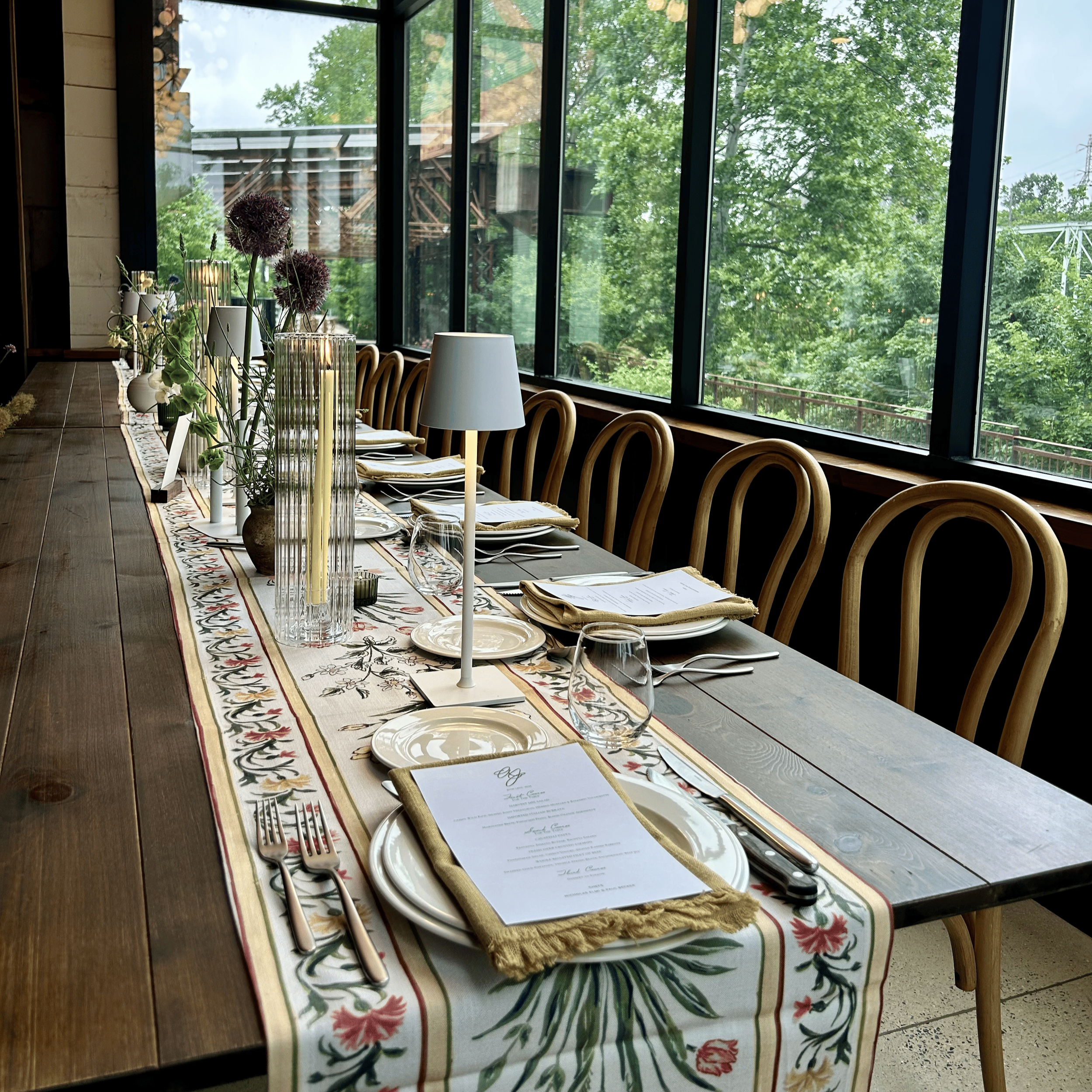A long wooden dining table set with plates, cutlery, wine glasses, and napkins, decorated with a patterned tablecloth and floral centerpieces, next to large windows with a view of green trees outside.