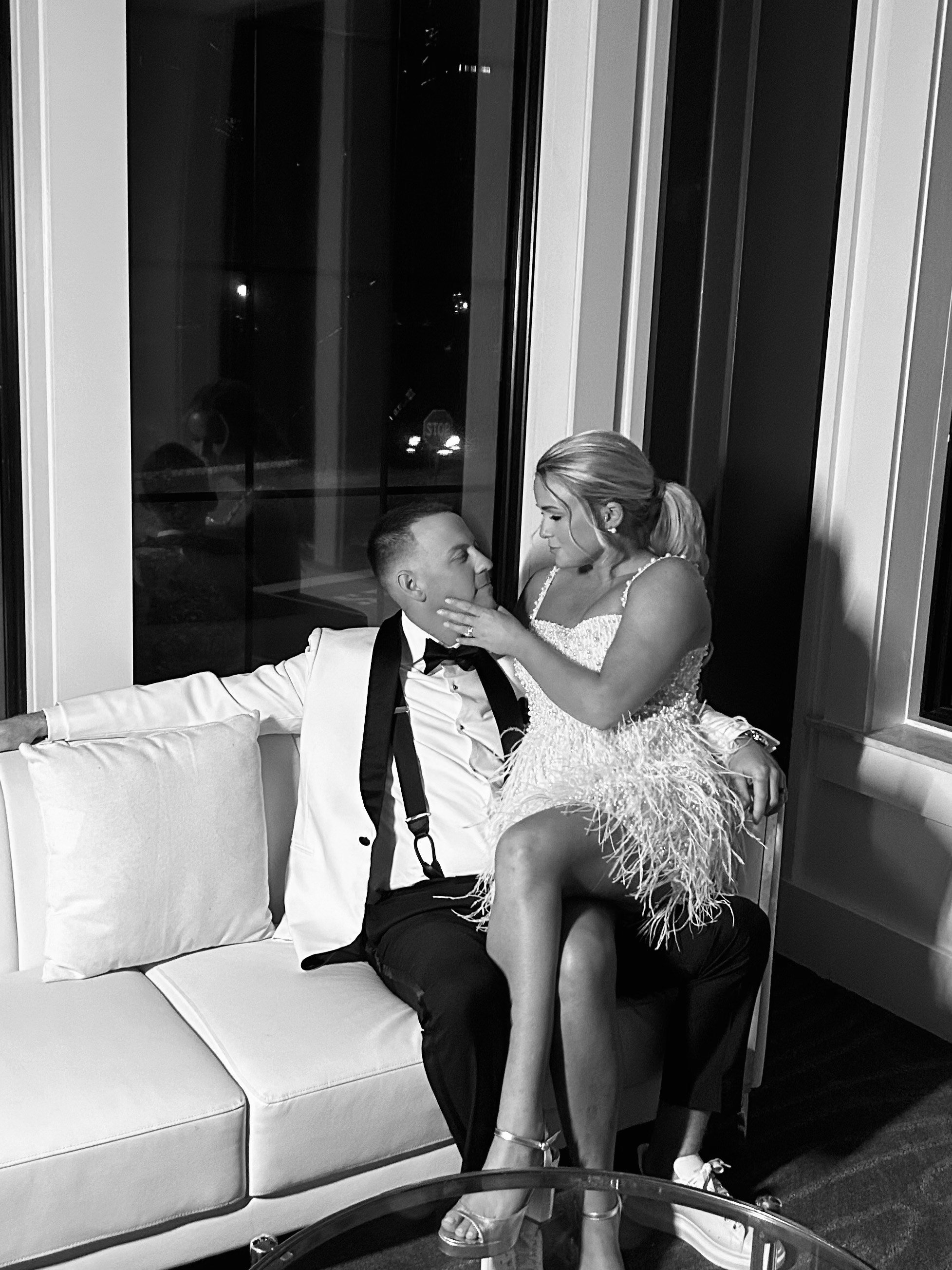 A black and white photo of a couple in formal attire sitting on a sofa in front of a window at night. The woman is sitting on the man's lap, touching his face, and both are looking into each other's eyes.