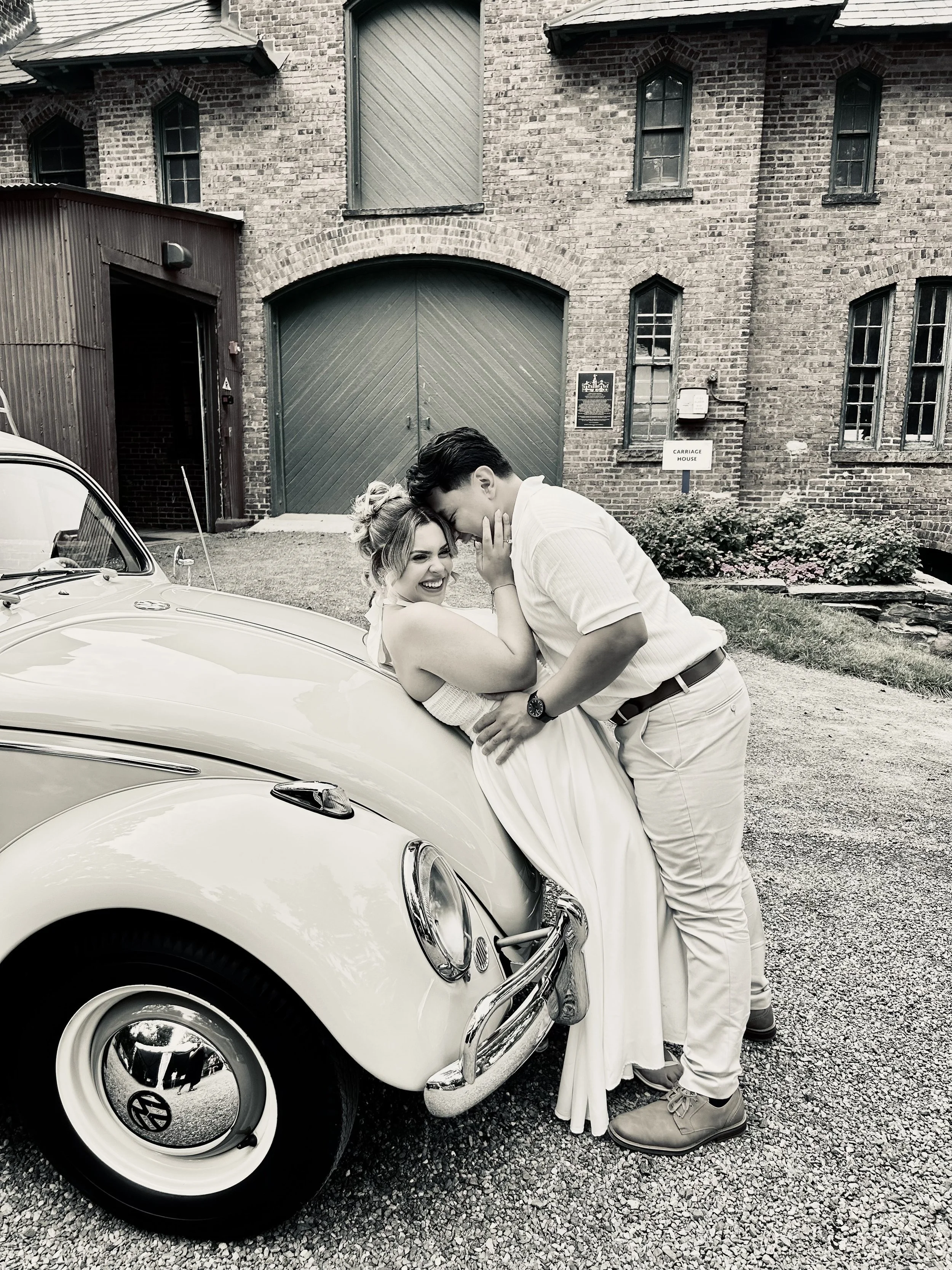 A couple shares a playful and intimate moment leaning against a vintage Volkswagen Beetle parked outside a brick building with a large wooden door and multiple windows. The woman is smiling and holding the man's face, while the man is leaning in close with his hand on her waist.