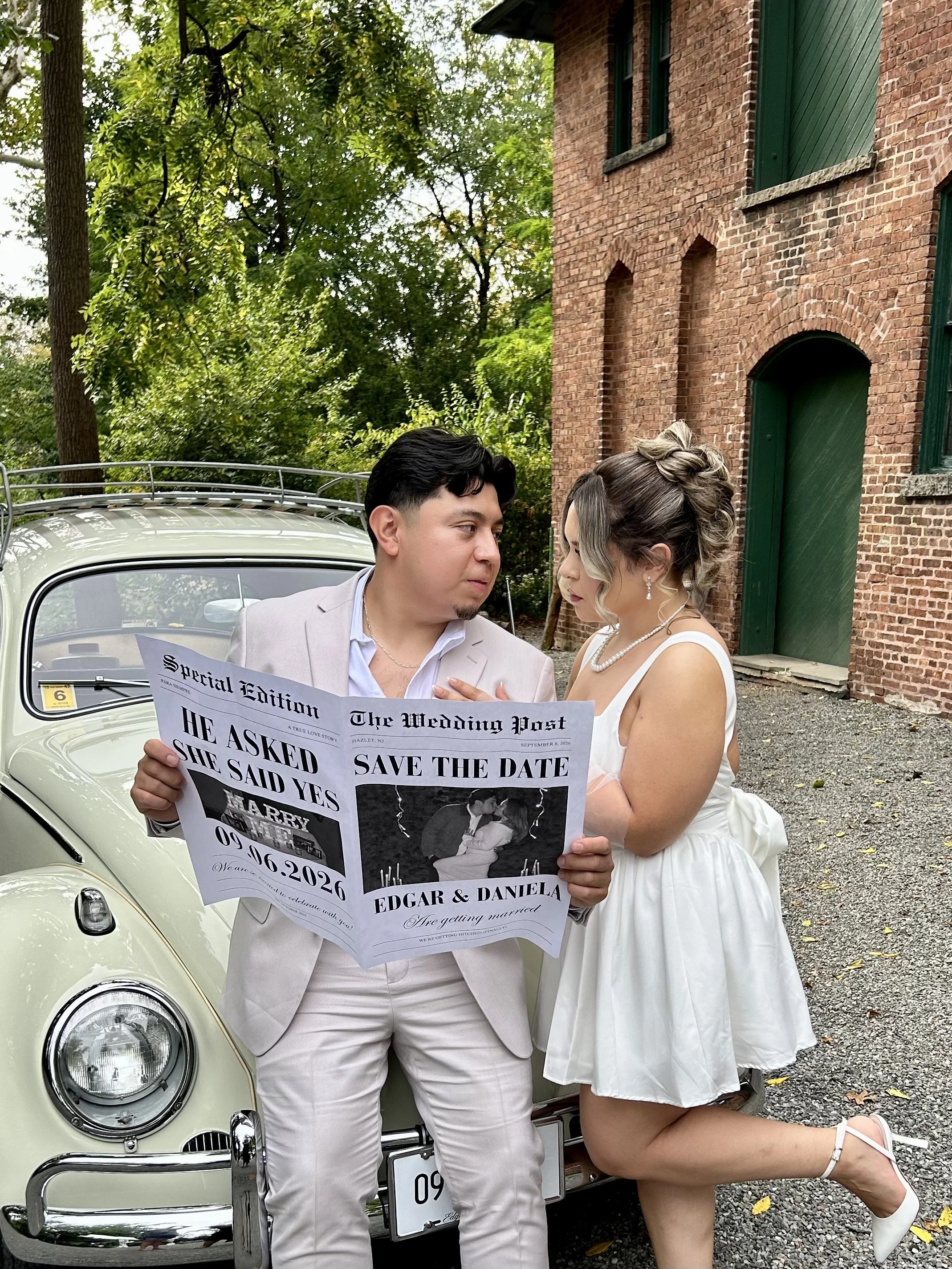 A man and woman dressed in wedding attire are standing outdoors next to a vintage car. The man is holding a newspaper with wedding announcements, looking at the woman, who is leaning on the car and looking at the man. The background features a brick building and green trees.