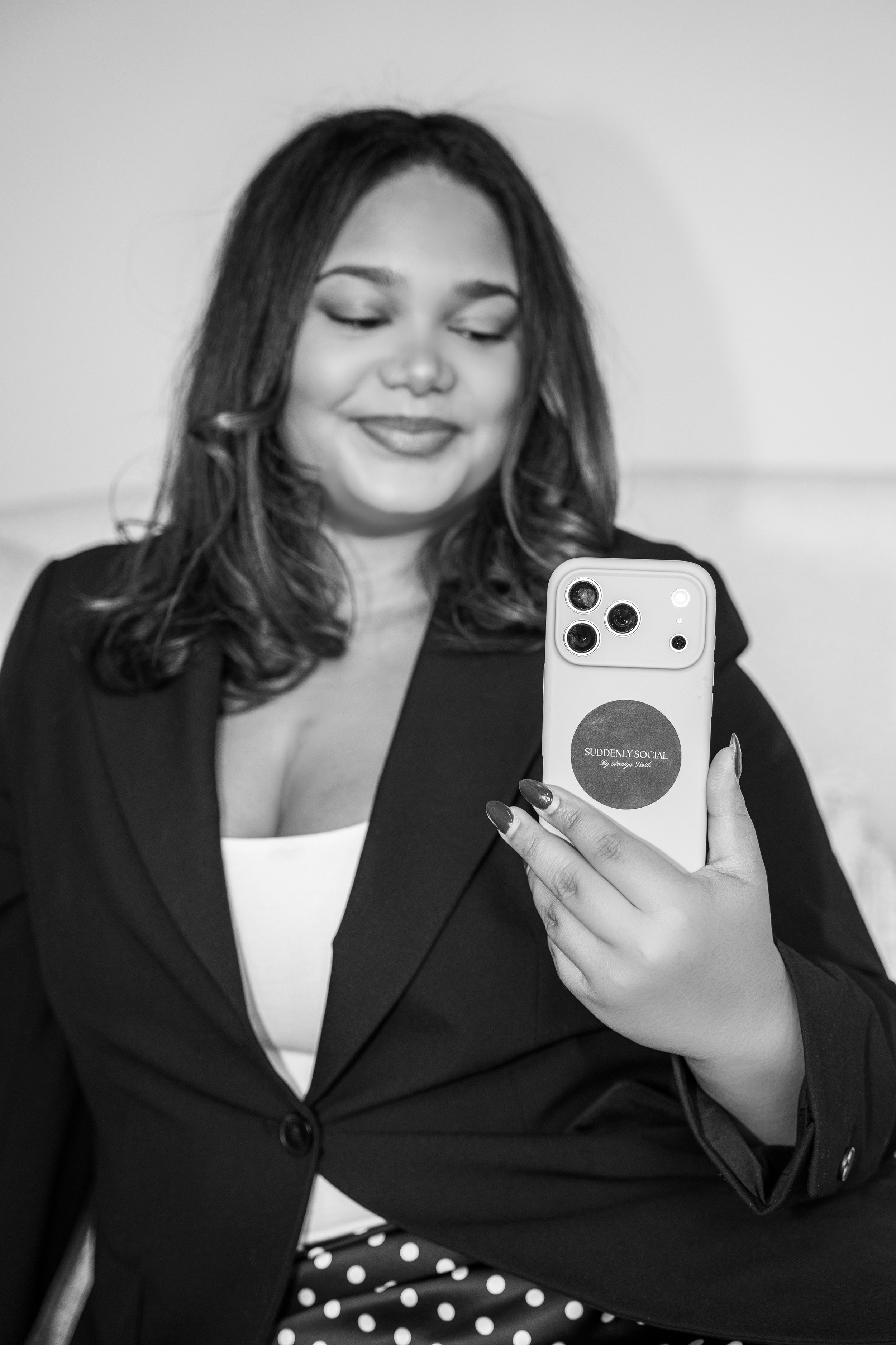 A woman taking a selfie with her smartphone, wearing a black blazer, white top, and polka dot skirt, with a smile and closed eyes.